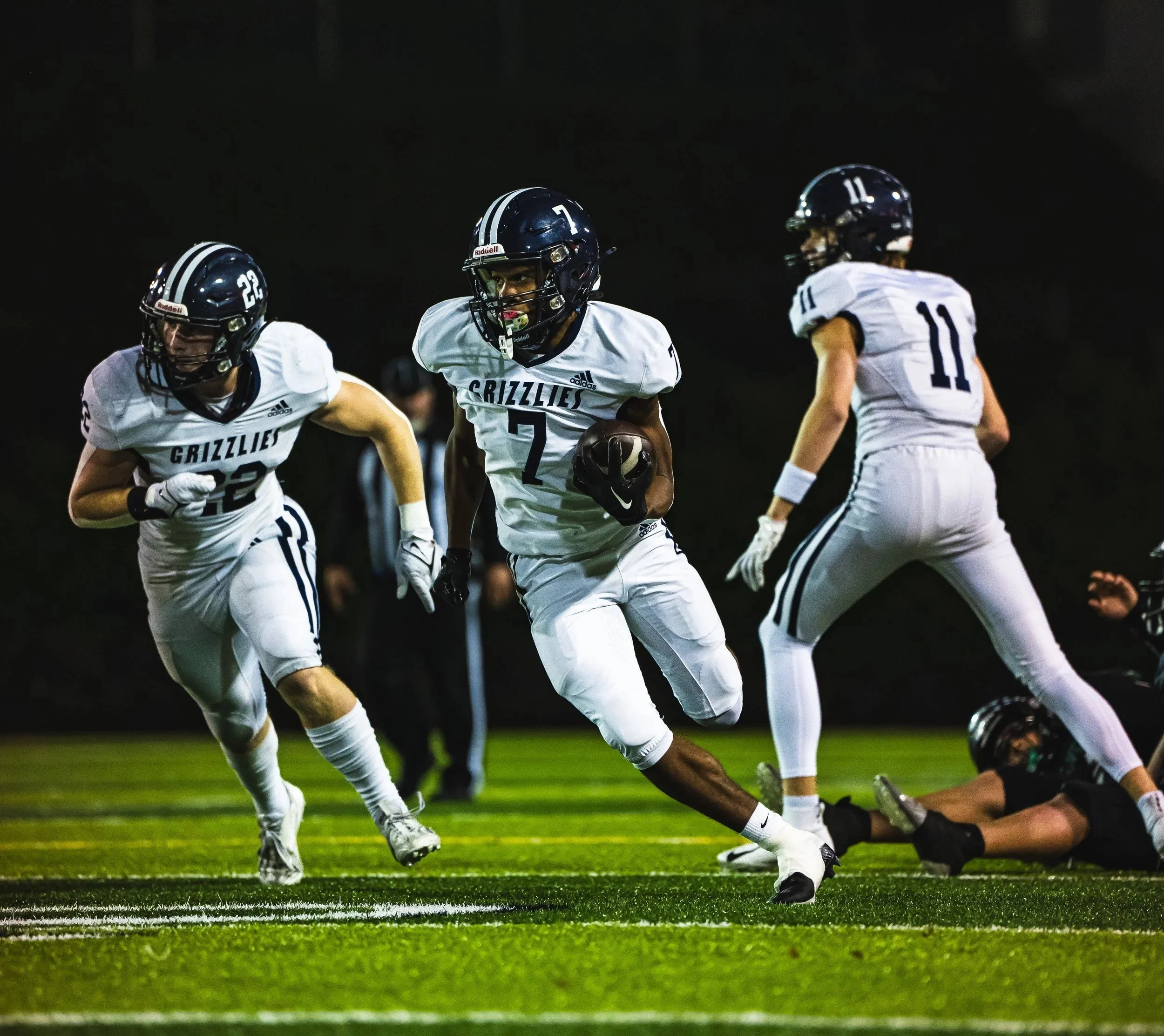 Football players in white uniforms and black helmets running on a field during a game at night.