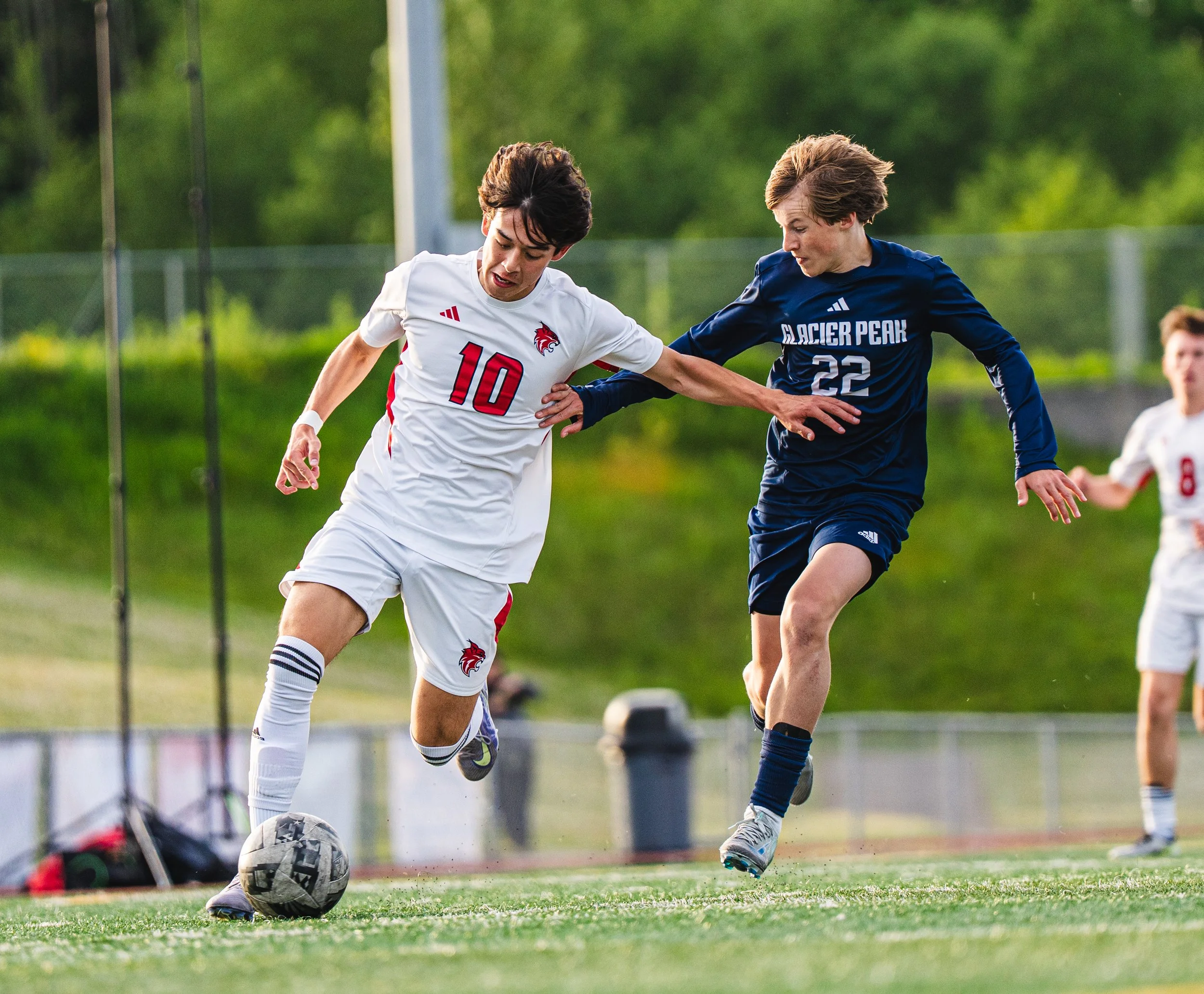 Two teenage boys playing soccer on a field, one in a white jersey with the number 10 and the other in a blue jersey with the number 22, competing for the ball during the daytime.