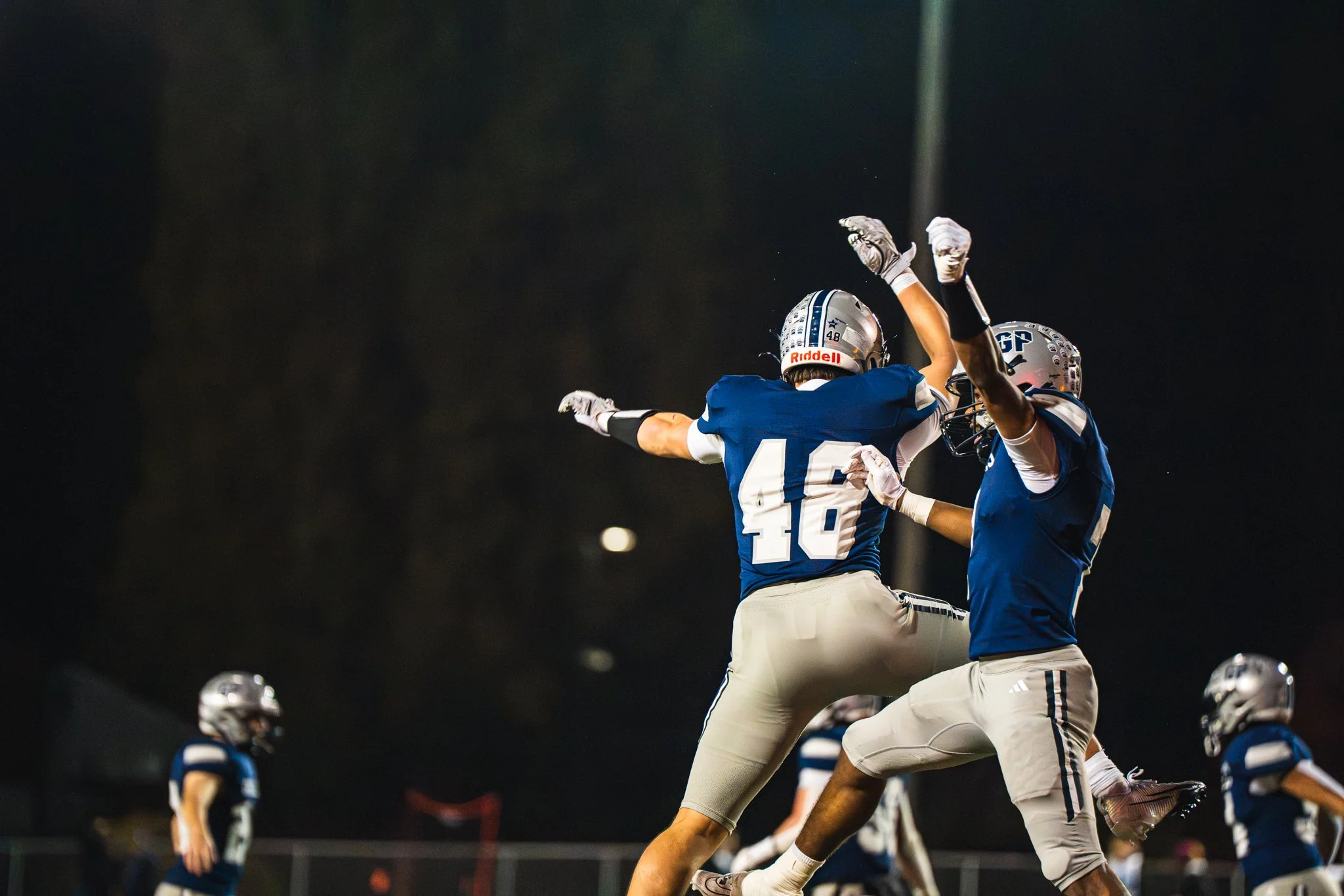 Football players in a blue and white uniform celebrating during a game at night, one with arms outstretched and another jumping with arms raised.