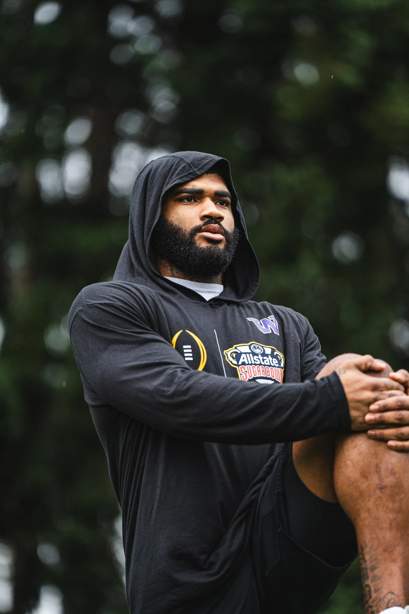 A man with a beard and dark hair wearing a black hoodie with the hood up, standing outdoors with a blurred background of trees, appears to be engaged in a physical activity or exercise.