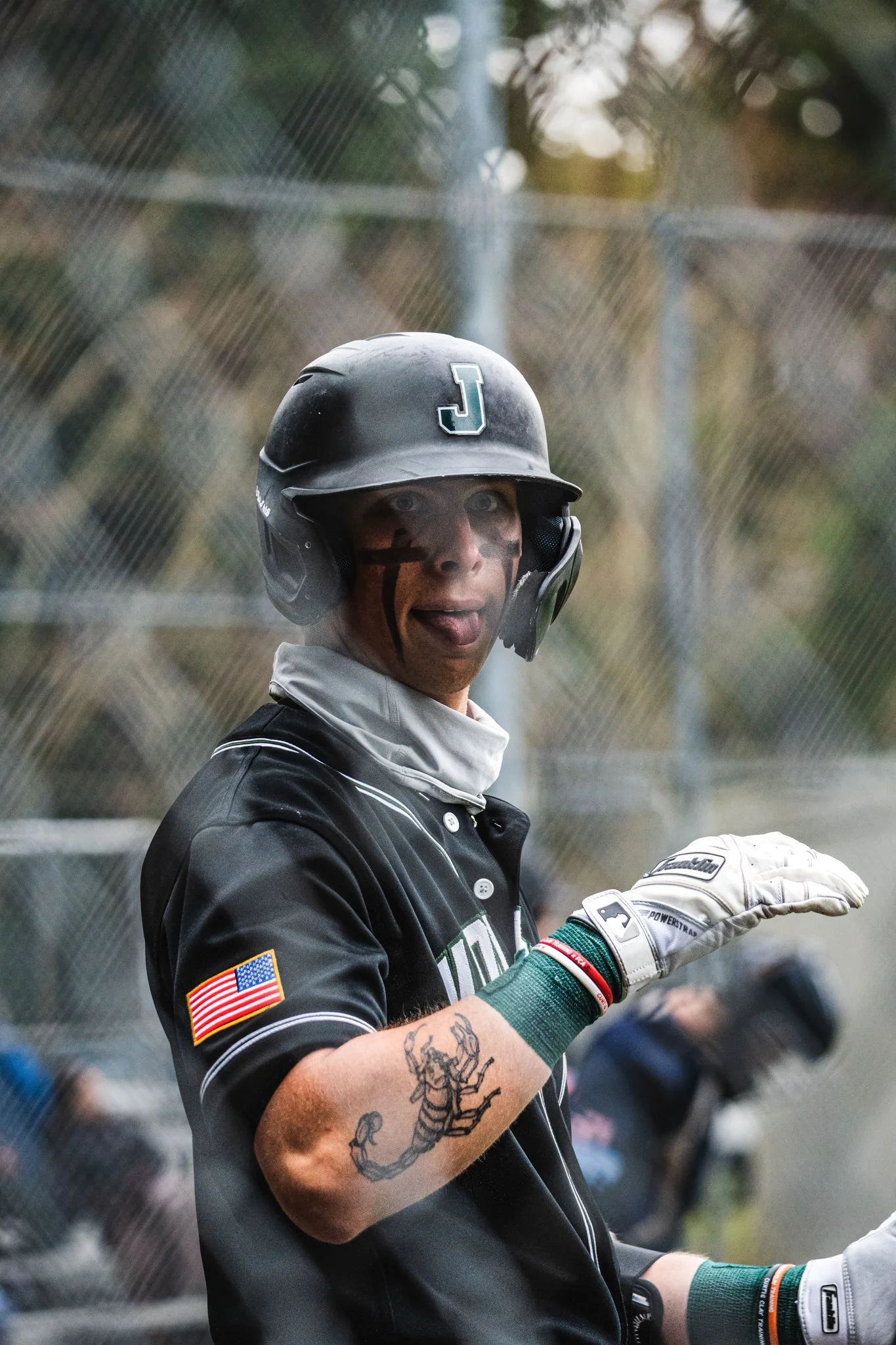 A person wearing a black helmet and black jacket, with tattoos on their arm, holding a baseball glove and standing near a chain-link fence during a baseball game or practice.