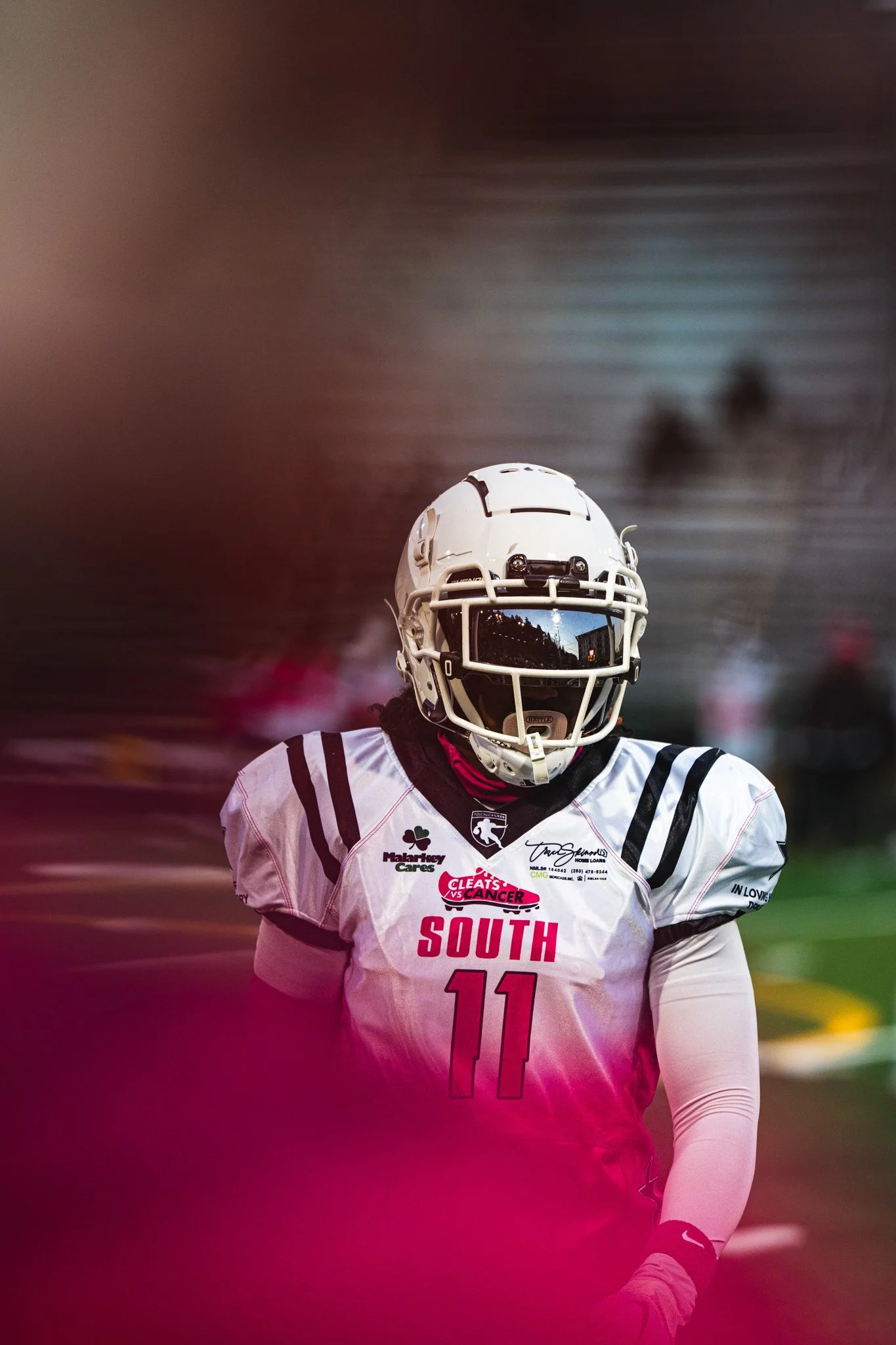 A football player wearing a white jersey with the number 11 and 'South' written on it, standing on the field with a football helmet, and a blurry background of a football stadium.