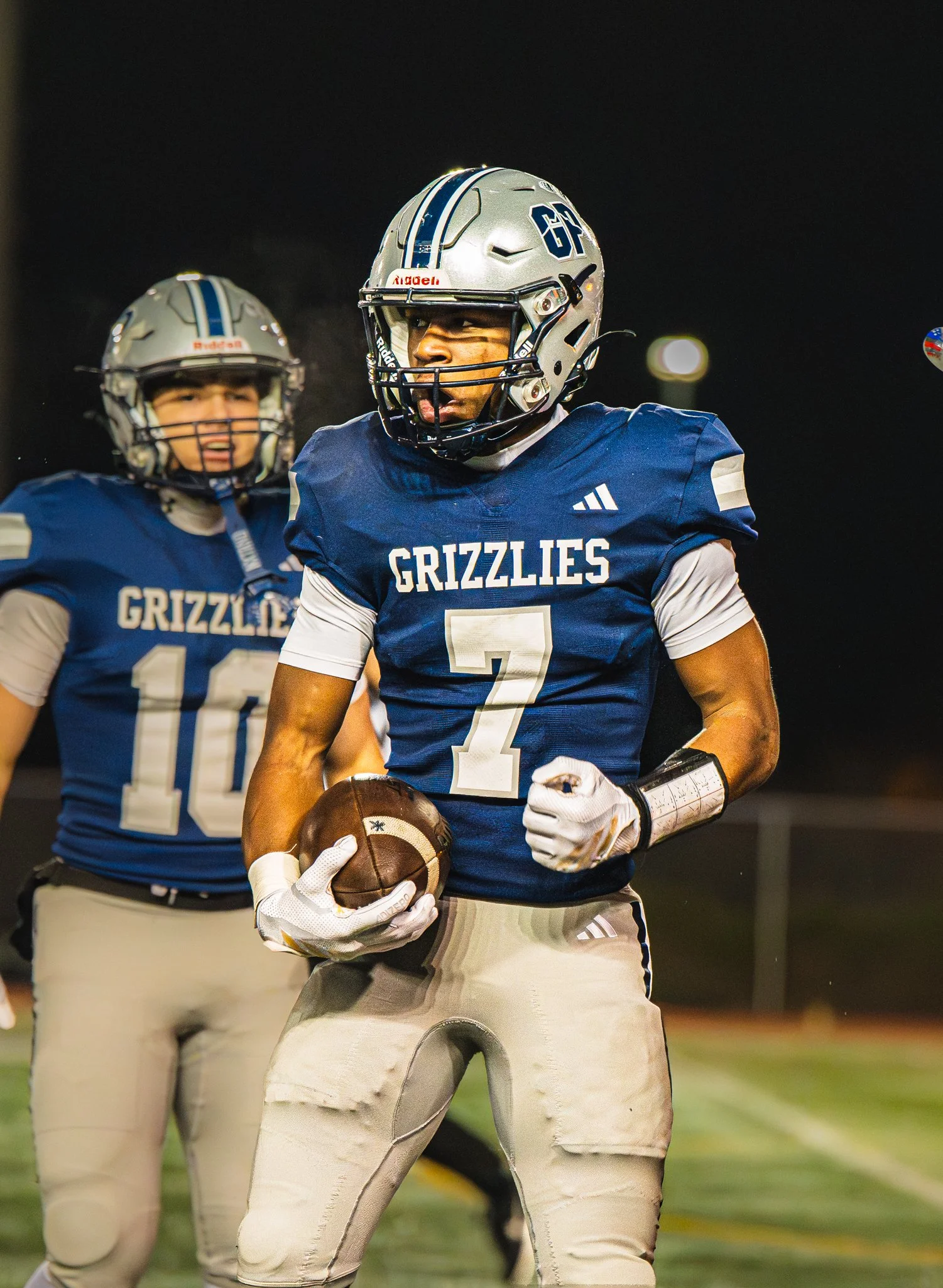 American football players wearing blue and white uniforms with ' GRIZZLIES ' written on their jerseys, one player holding a football, on a field at night.