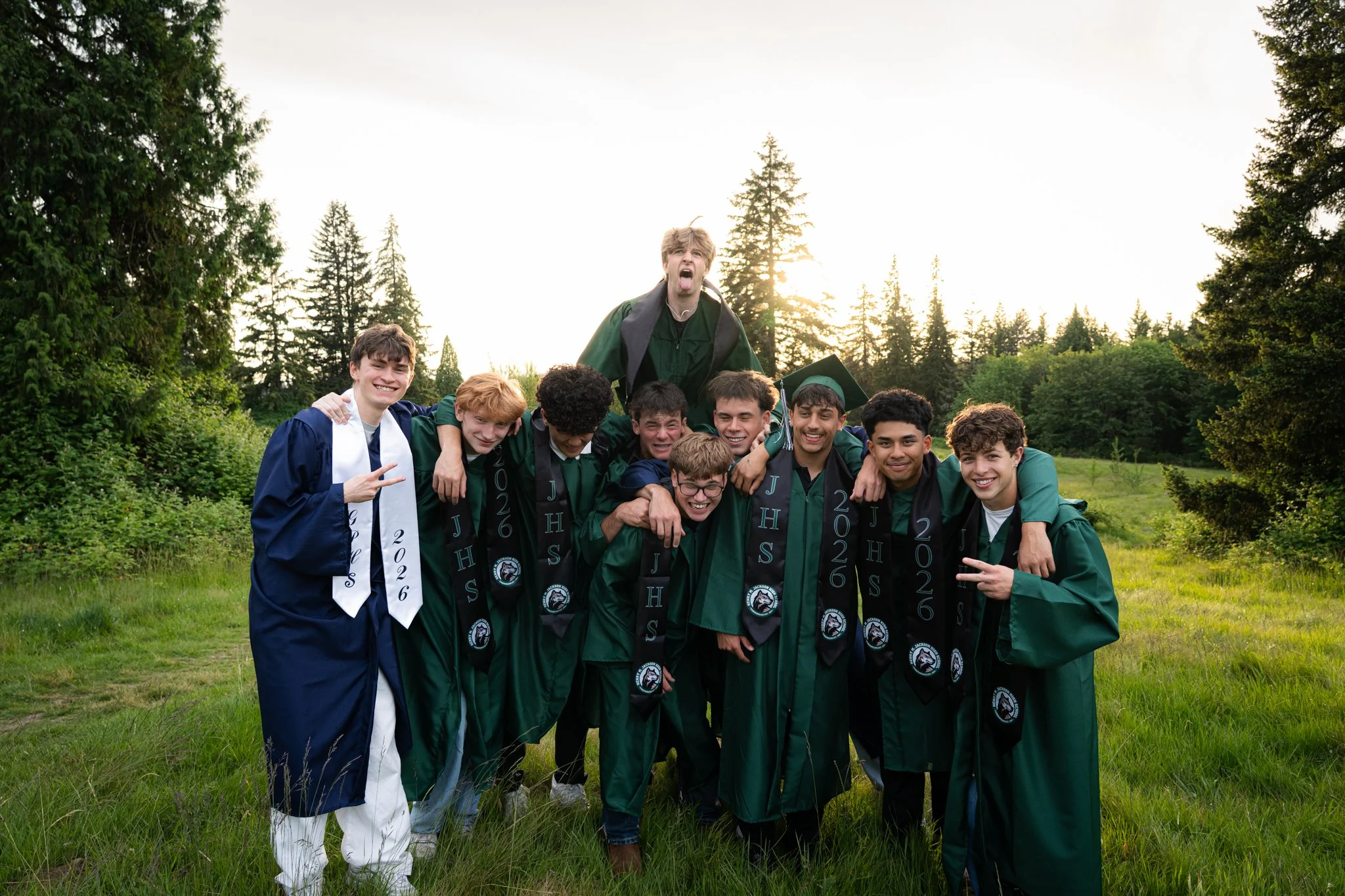 Group of smiling young men in graduation caps and gowns posing outdoors in a green field with trees and sunlight in the background.