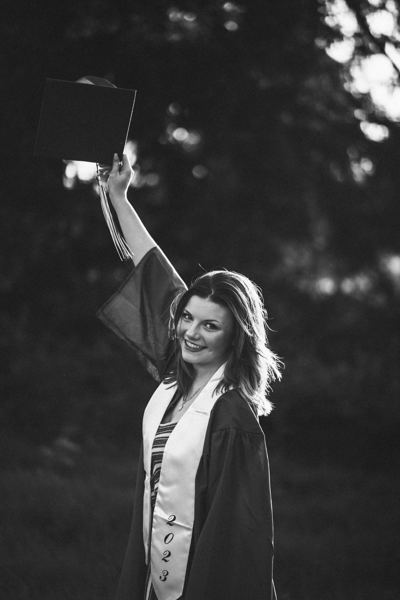 A young woman in a graduation gown and cap, smiling, holding up her cap, standing outdoors with blurred trees in the background, wearing a sash with the year 2023.