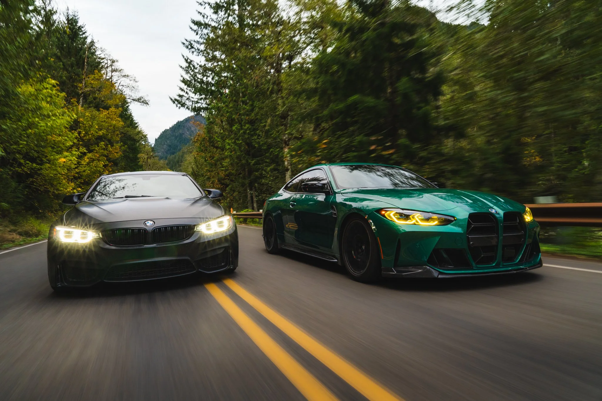 Two sports cars driving on a winding mountain road surrounded by green trees, with mountains in the background and a cloudy sky.