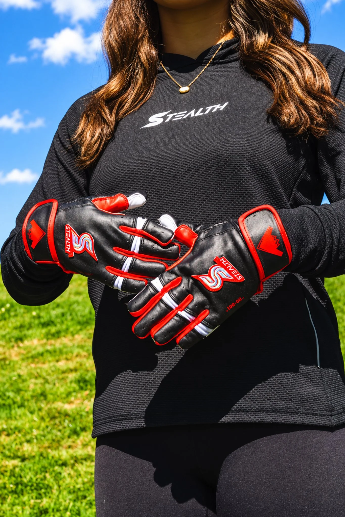 A woman in athletic clothing wearing black and red goalkeeper gloves, a black long-sleeve shirt with the word 'STEALTH' on it, a gold necklace, and standing against a background of grass, blue sky, and clouds.