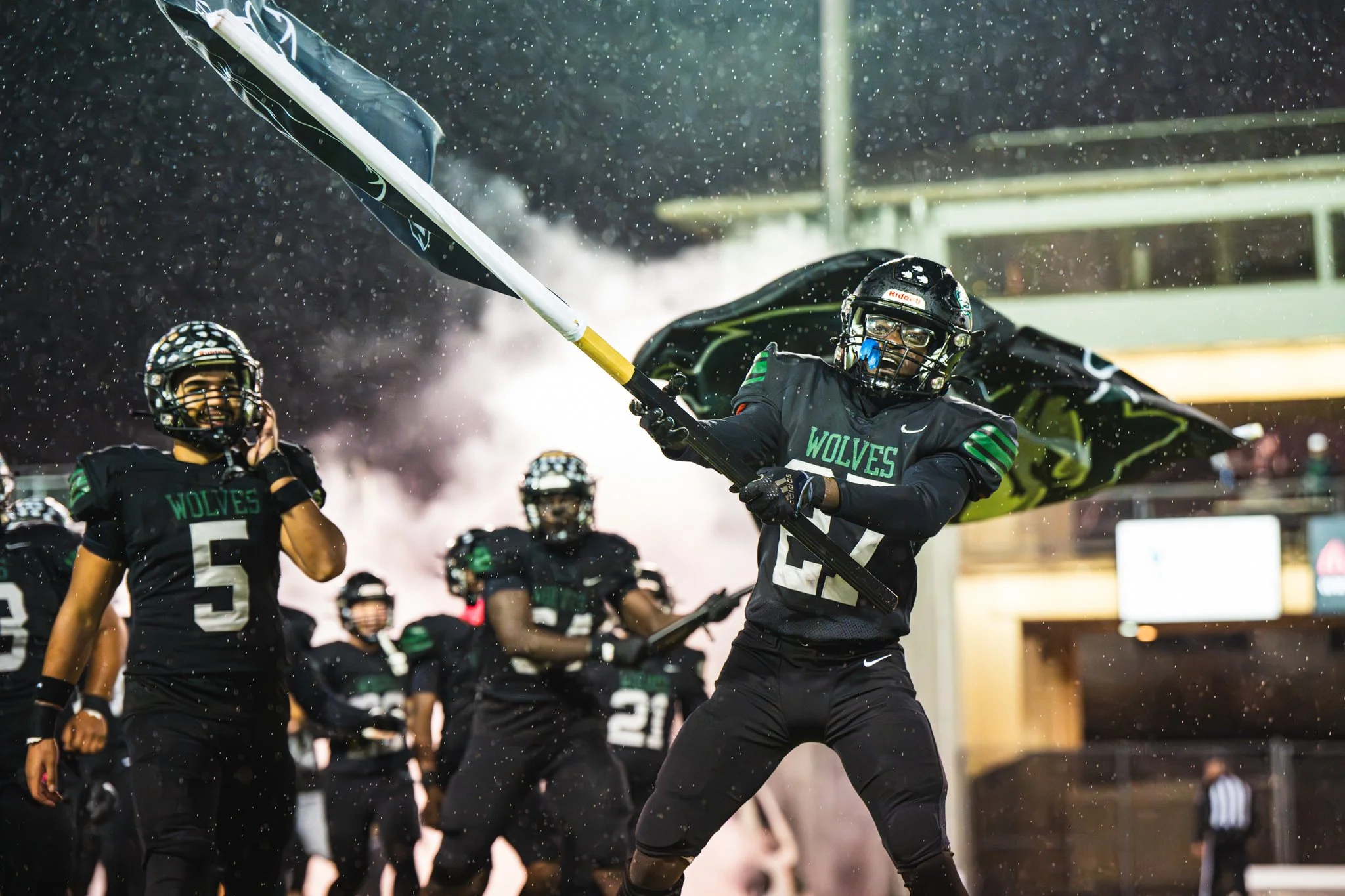 Football players in black uniforms with green accents, helmets, and face masks celebrating on the field, with one player raising a flag.