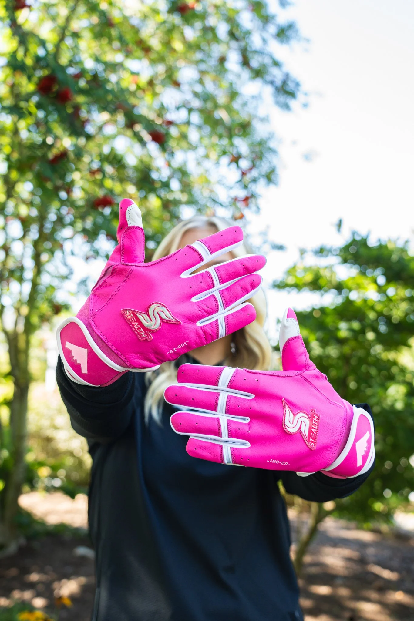 Person wearing bright pink gardening gloves, standing outdoors with trees and sunlight in the background, making a gesture with hands.