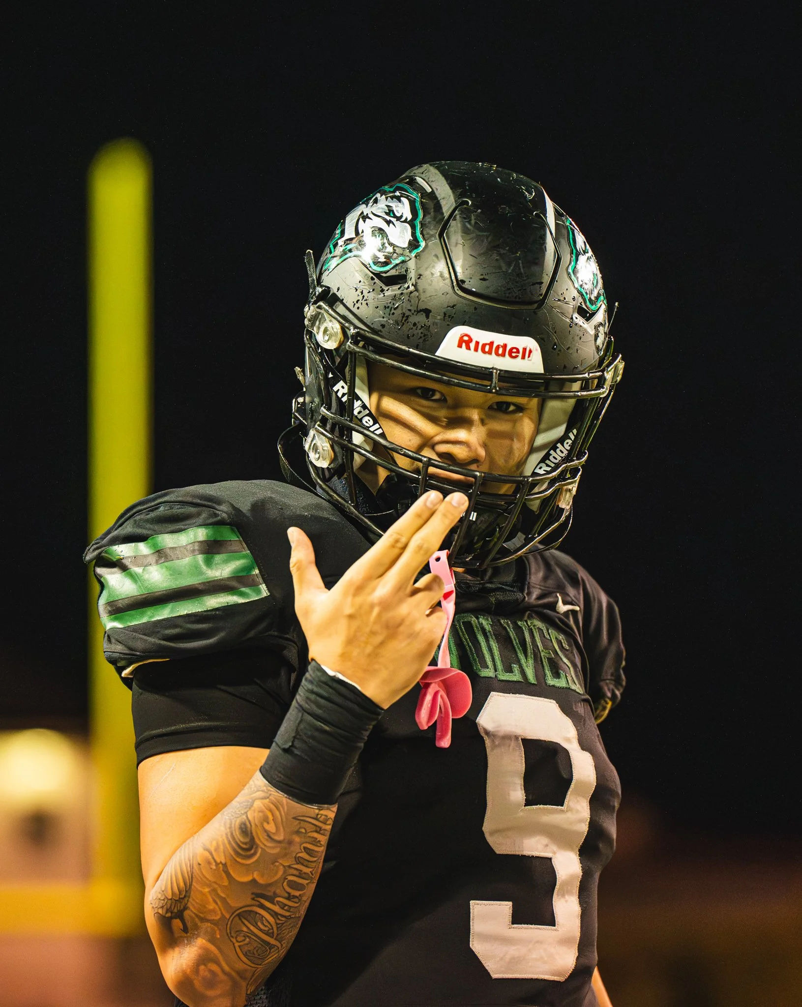 Close-up of an American football player wearing a black and green uniform and a black helmet with a logo, making a hand gesture on the field at night.