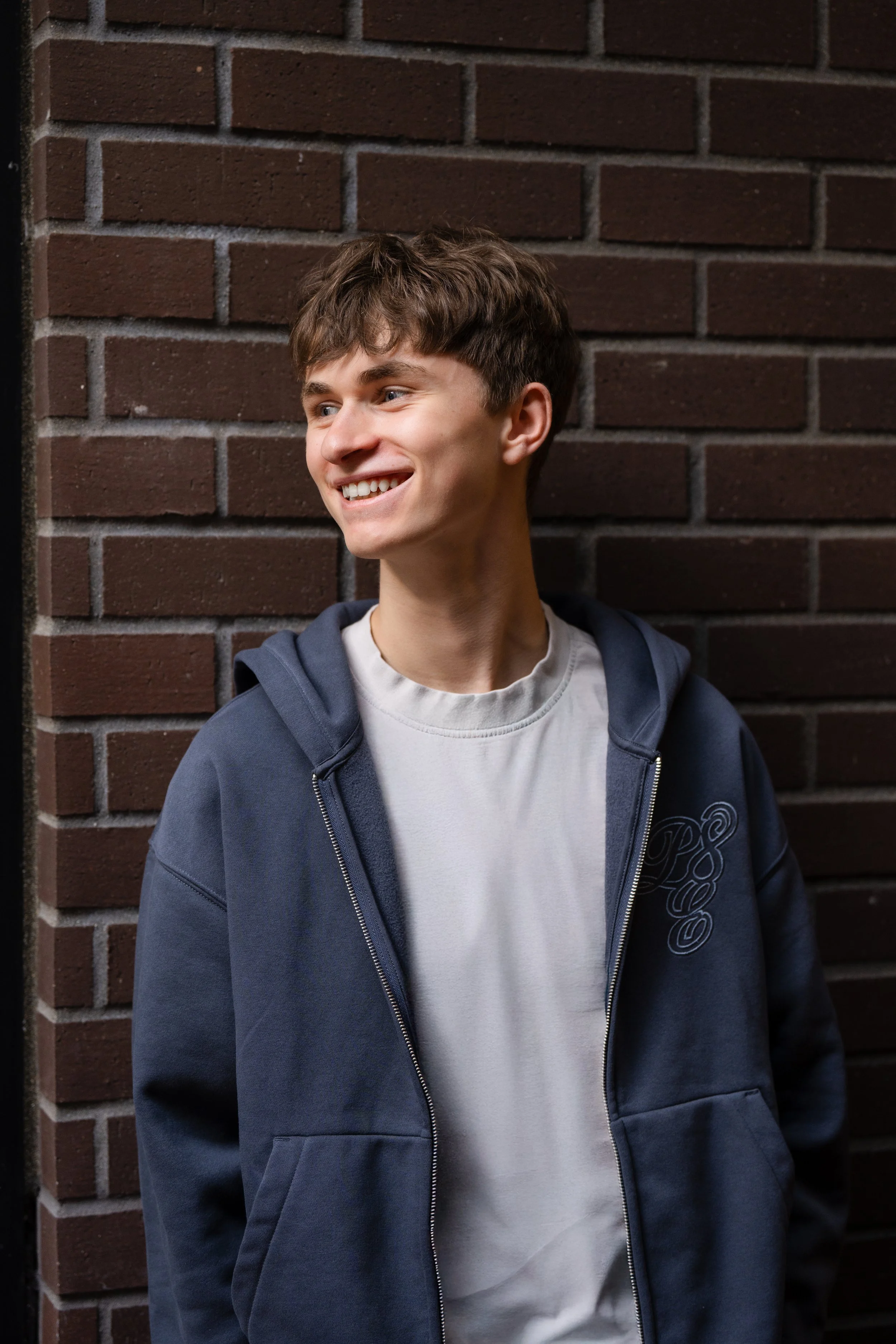 A young man with brown hair smiling while leaning against a brick wall, wearing a white T-shirt and a blue hoodie.