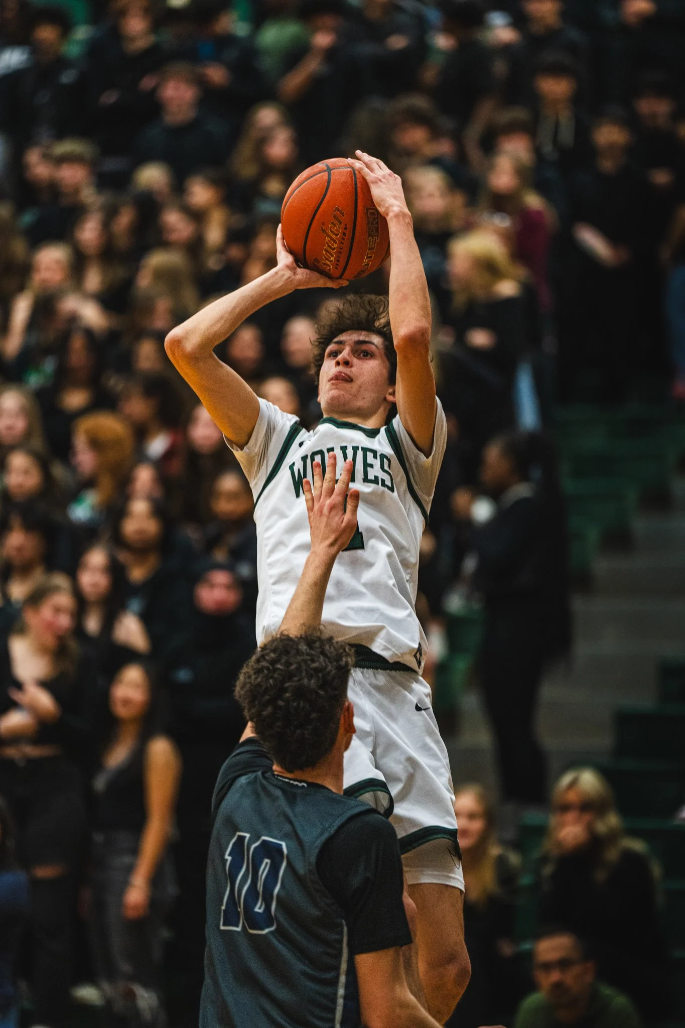 A basketball game scene with a player in a white jersey jumping to shoot while another player in a dark jersey tries to block, with an audience in the background.