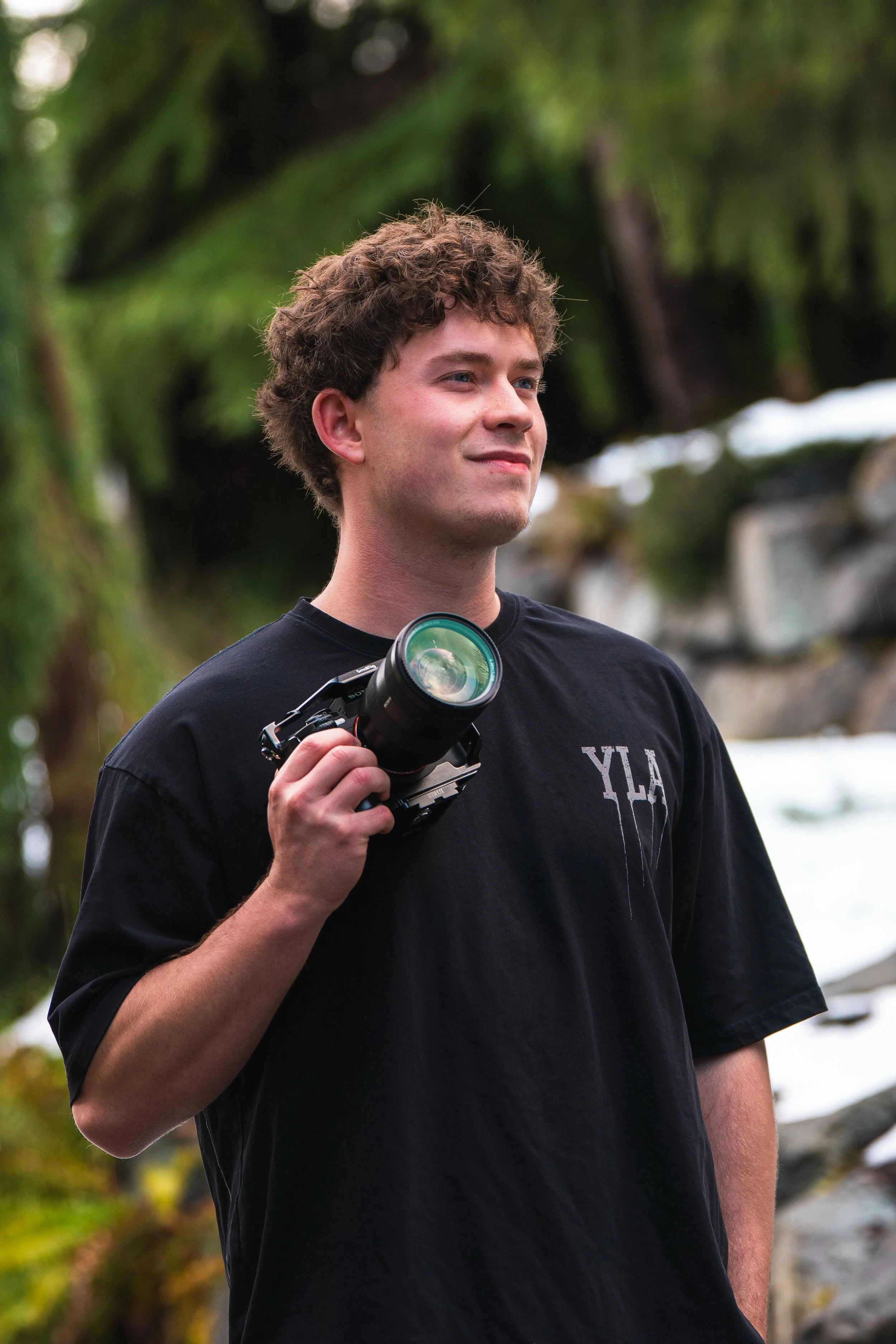 A young man with curly hair holding a camera strap over his shoulder, standing outdoors in a lush, natural setting.