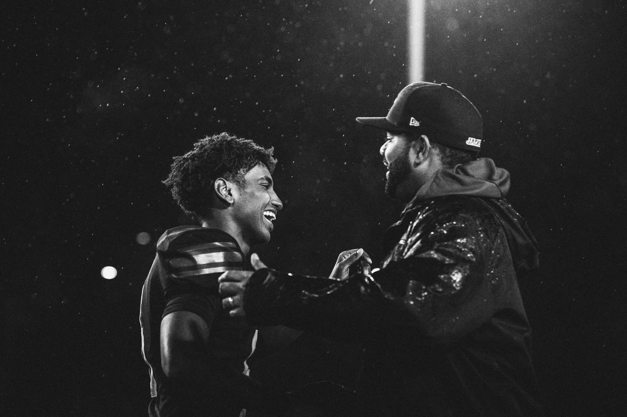 Two people, a woman with short curly hair and a man with a beard and cap, smiling and holding hands in the rain at night.