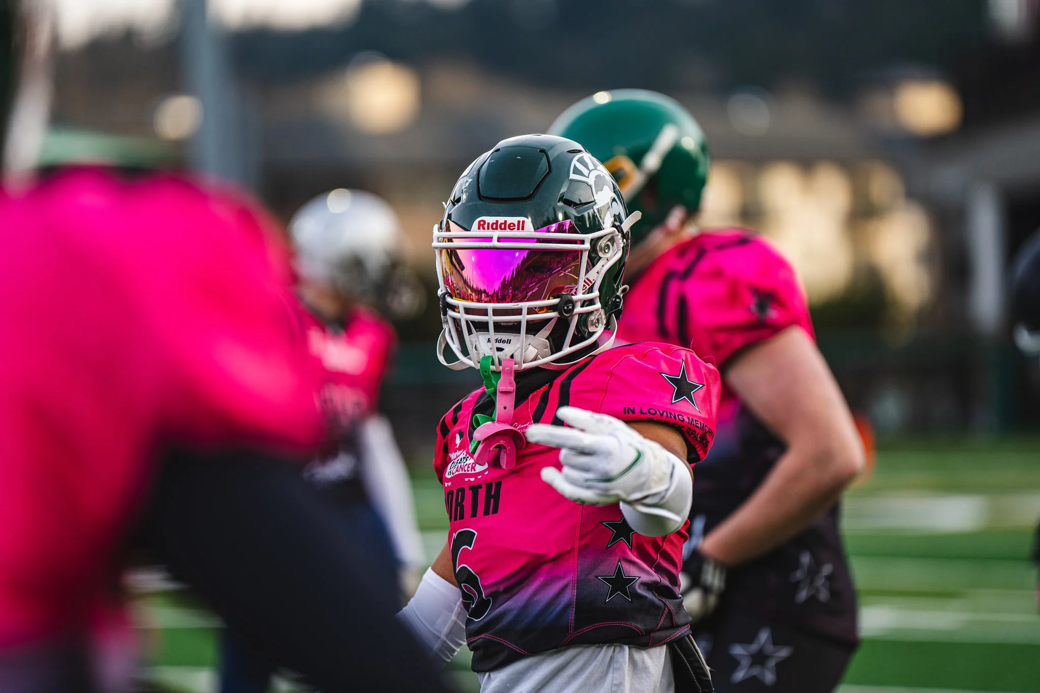 A young football player in a pink jersey with black stars, wearing a helmet with a pink tinted visor, is holding a football and making a gesture with her hand during a game or practice.