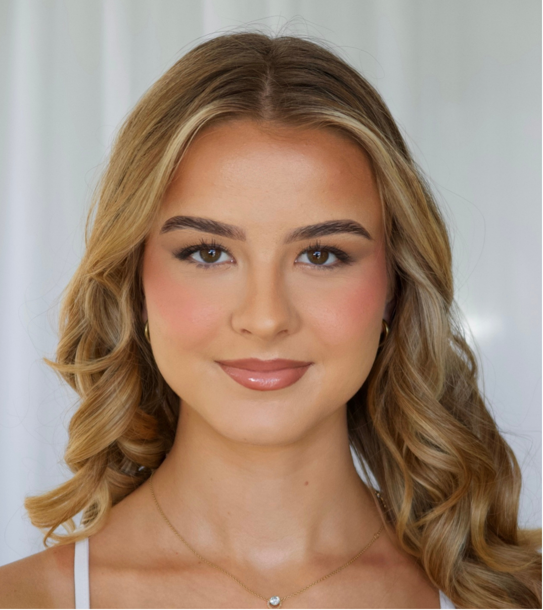 Close-up of a young woman with wavy blonde hair, brown eyes, wearing makeup, gold earrings, and a delicate necklace, smiling softly at the camera.