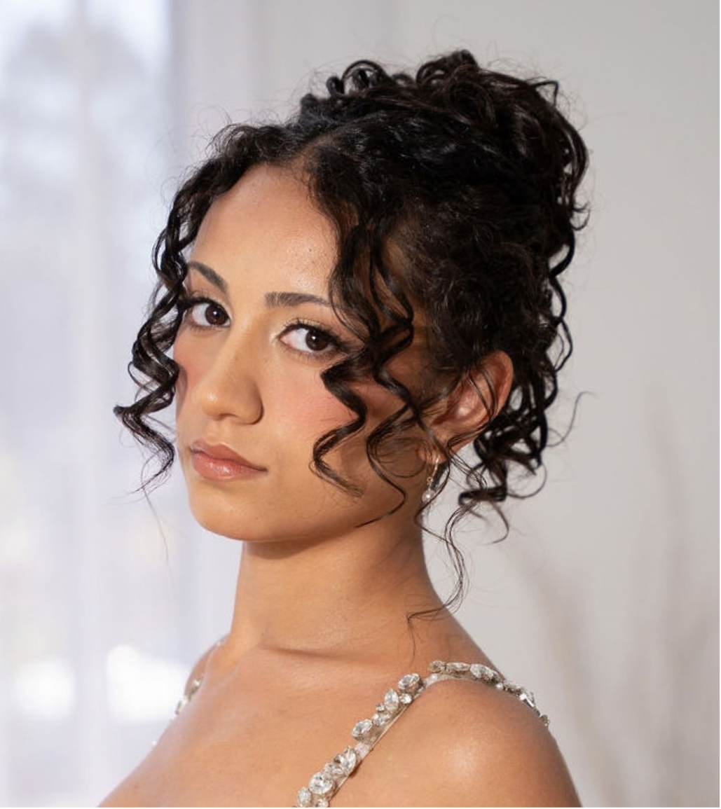Close-up of a woman with dark curly hair, wearing makeup, and a dress with rhinestone straps.