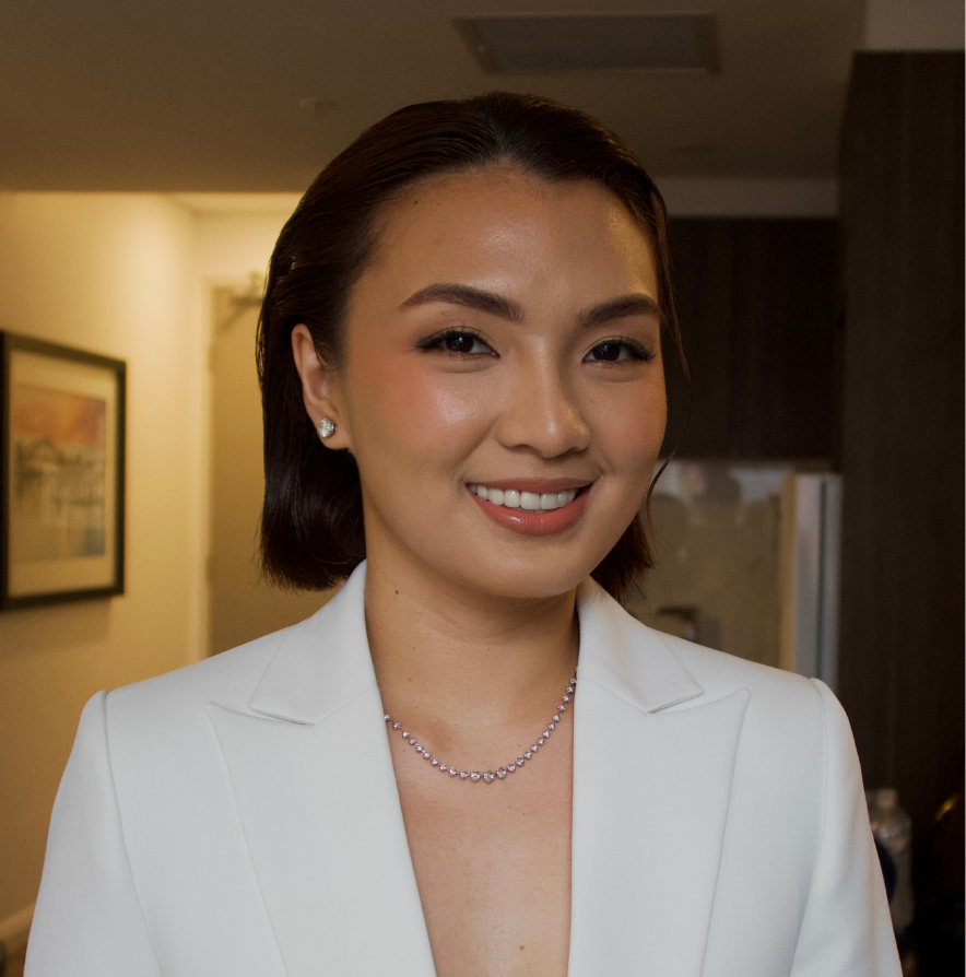 A woman with short dark hair, dressed in a white blazer, smiling and wearing a diamond necklace and earrings inside a well-lit room.