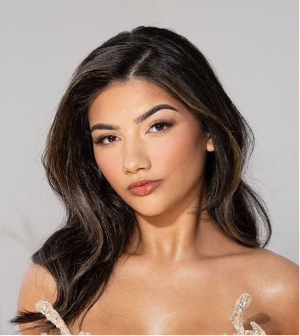 Close-up of a woman with long, wavy dark hair, light makeup, and a neutral background.