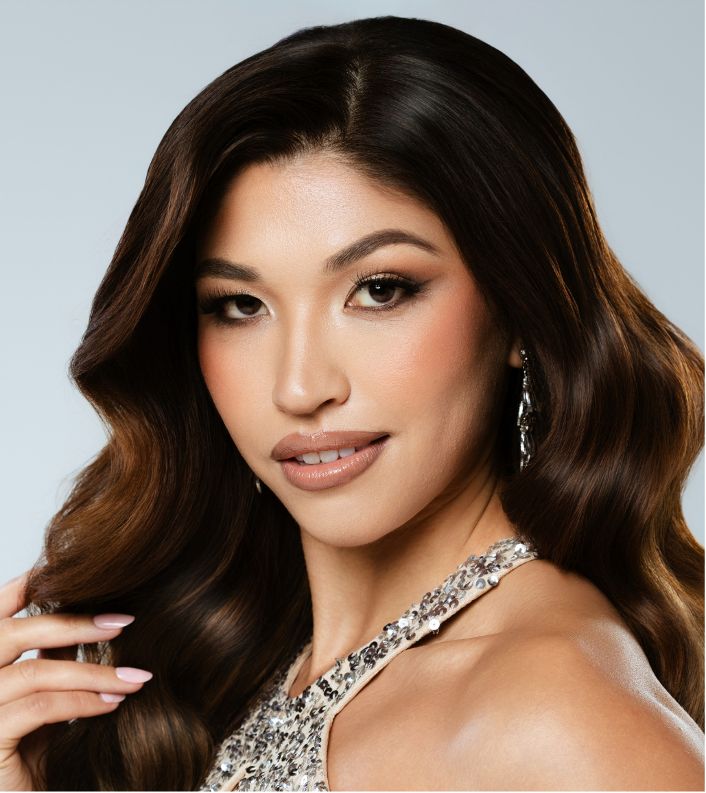A woman with long, wavy dark brown hair, wearing elegant makeup and a beaded dress, looking at the camera with a slight smile.