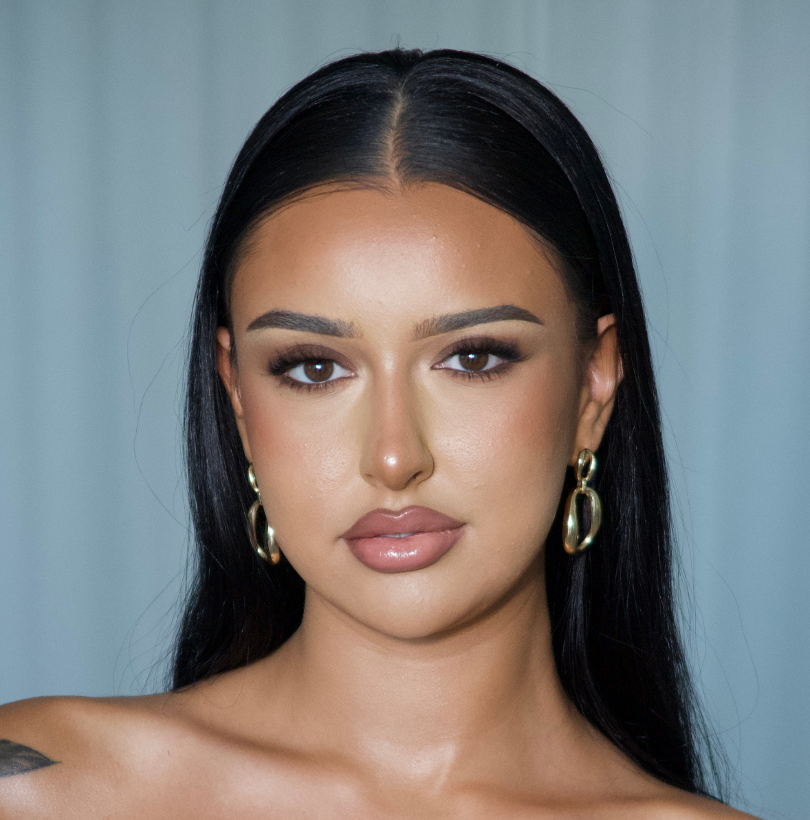 Close-up portrait of a woman with long black hair, brown eyes, and gold hoop earrings, wearing makeup, looking directly at the camera.