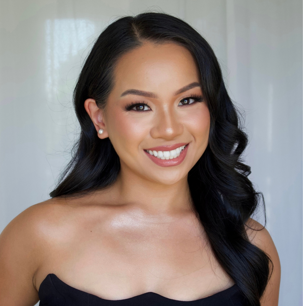 A smiling woman with long black hair, wearing pearl earrings and a strapless black top, standing in front of a light-colored background.
