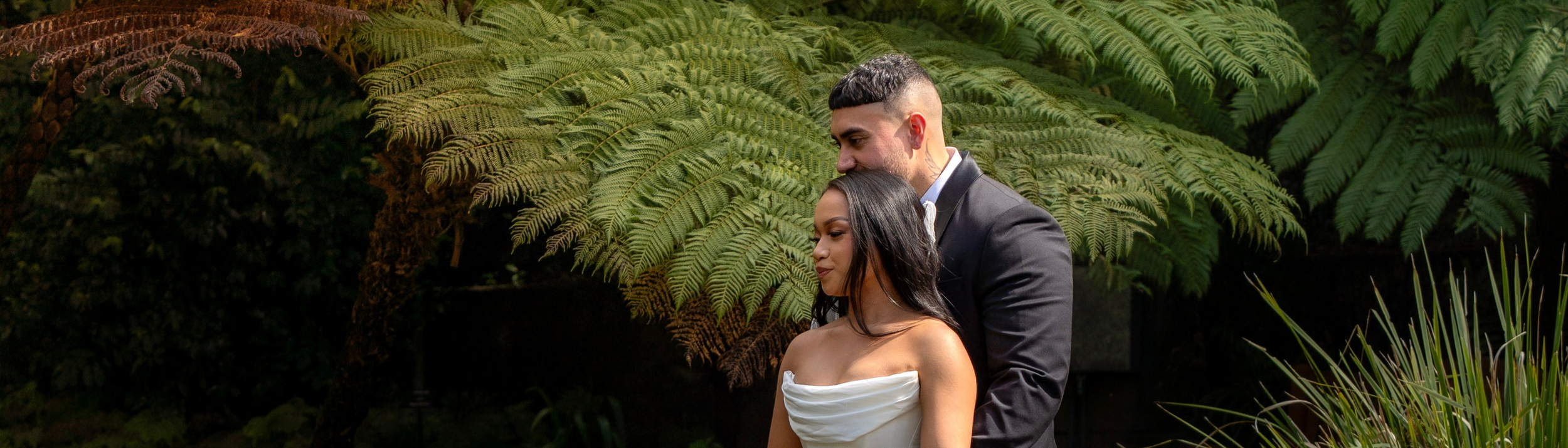 A man and woman standing close together outdoors in front of lush green ferns, with the man embracing the woman from behind.