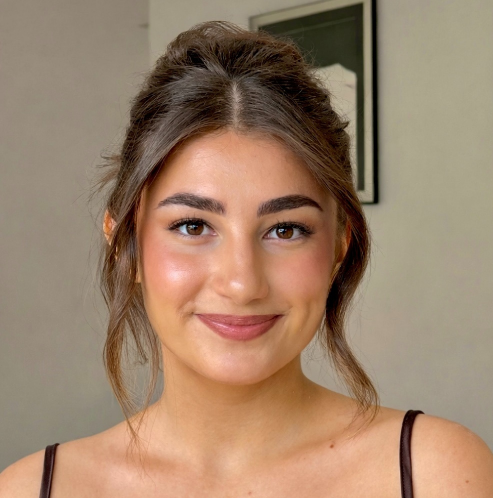 A young woman with brown hair styled in an updo, smiling at the camera, wearing a black spaghetti strap top, with a framed photograph behind her on a wall.