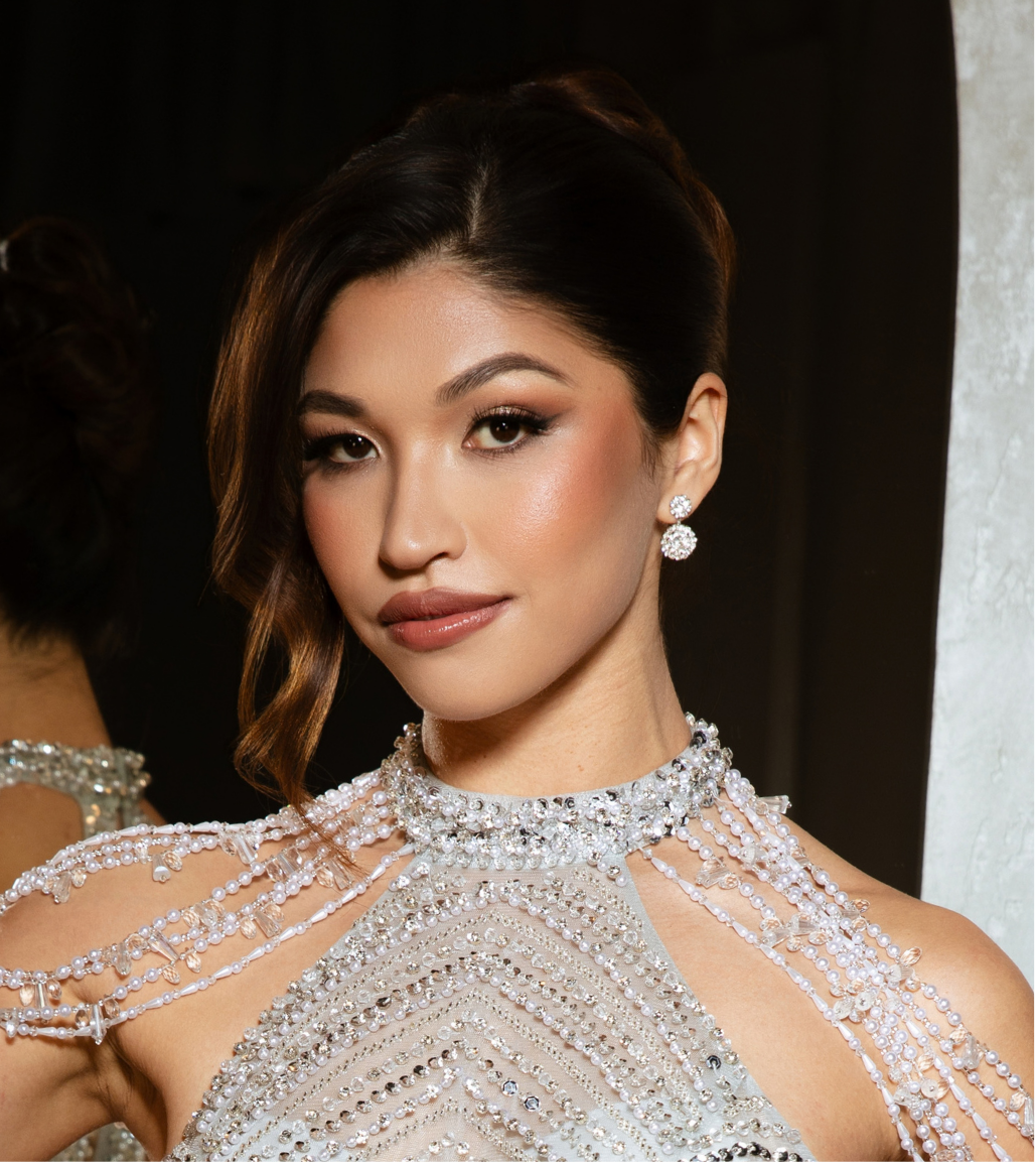 A woman with dark, wavy hair styled to one side, wearing elegant makeup, diamond earrings, and a detailed pearl and rhinestone dress.