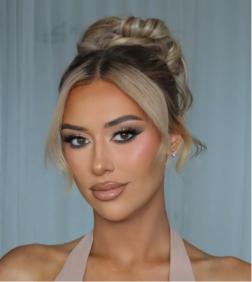 Close-up of a young woman with blonde hair styled in an updo, wearing makeup with bold eye makeup and nude lipstick, and a beige top, against a neutral background.
