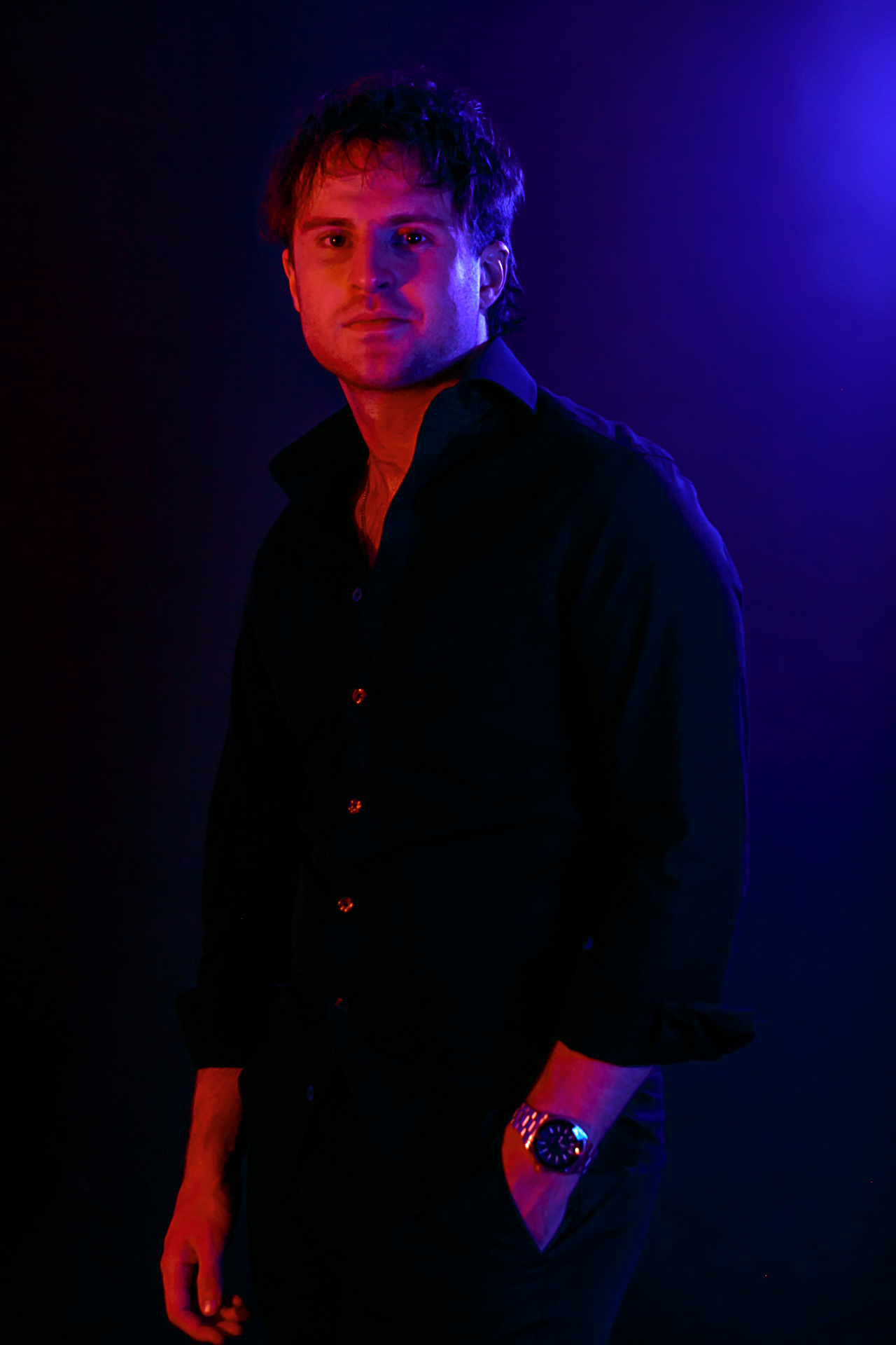 DJ Sona with dark hair and light skin, wearing a black button-up shirt and a silver watch, standing against a dark background with purple and blue lighting.