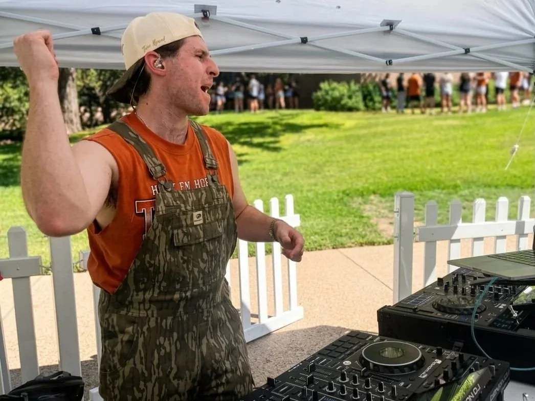 DJ Sona at an outdoor tailgate event, wearing camouflage overalls over an orange tank top, a beige baseball cap, and in front of DJ equipment, with a crowd of people in the background.