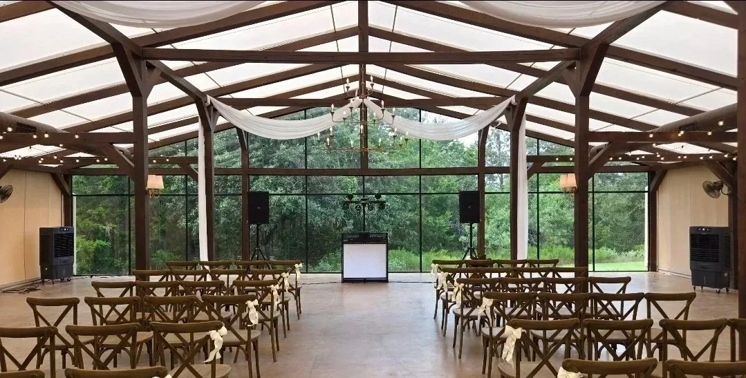 Indoor wedding ceremony venue with wooden chairs decorated with white ribbons, large glass walls showing greenery outside, and a DJ table at the front, decorated with drapes and lights.