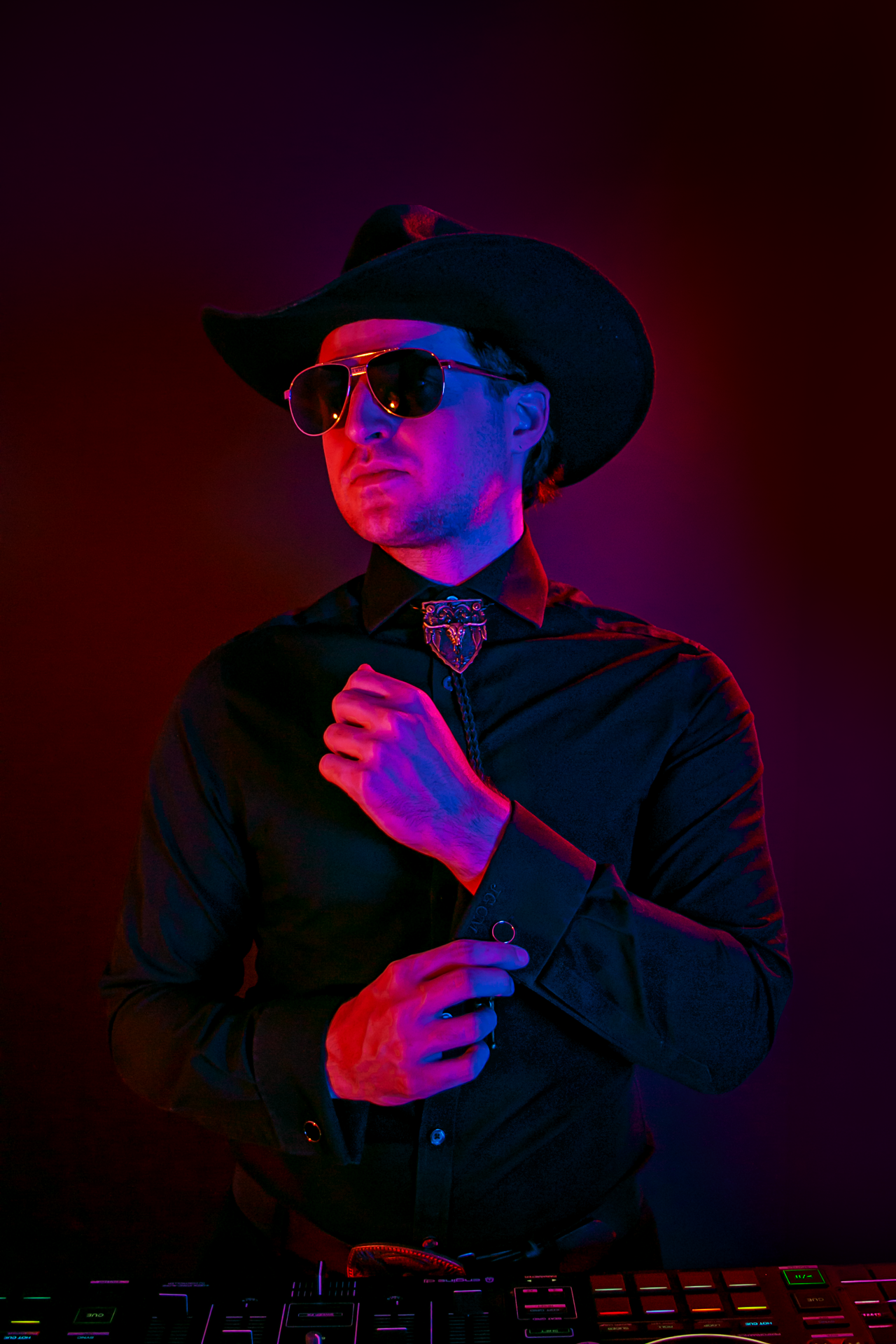 DJ Sona dressed in black wearing a wide-brimmed hat, sunglasses, and a bolo tie, standing in front of a colorful, neon-lit background.