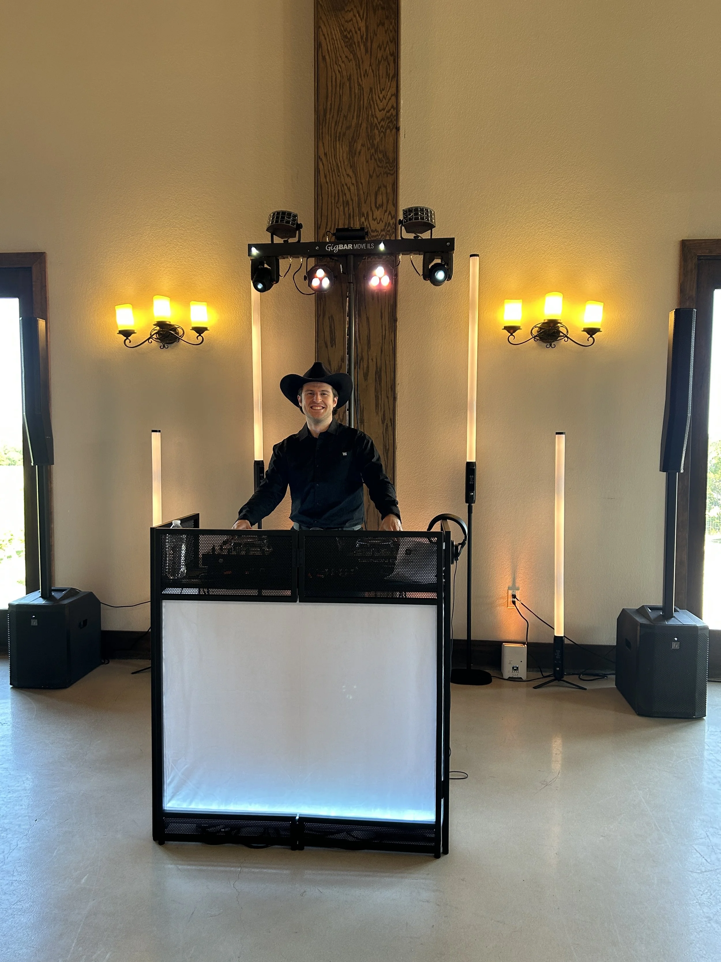 DJ Sona wearing a black cowboy hat and black shirt smiling while standing behind DJ equipment at an indoor wedding with yellow wall sconces and a DJ lighting rig overhead.