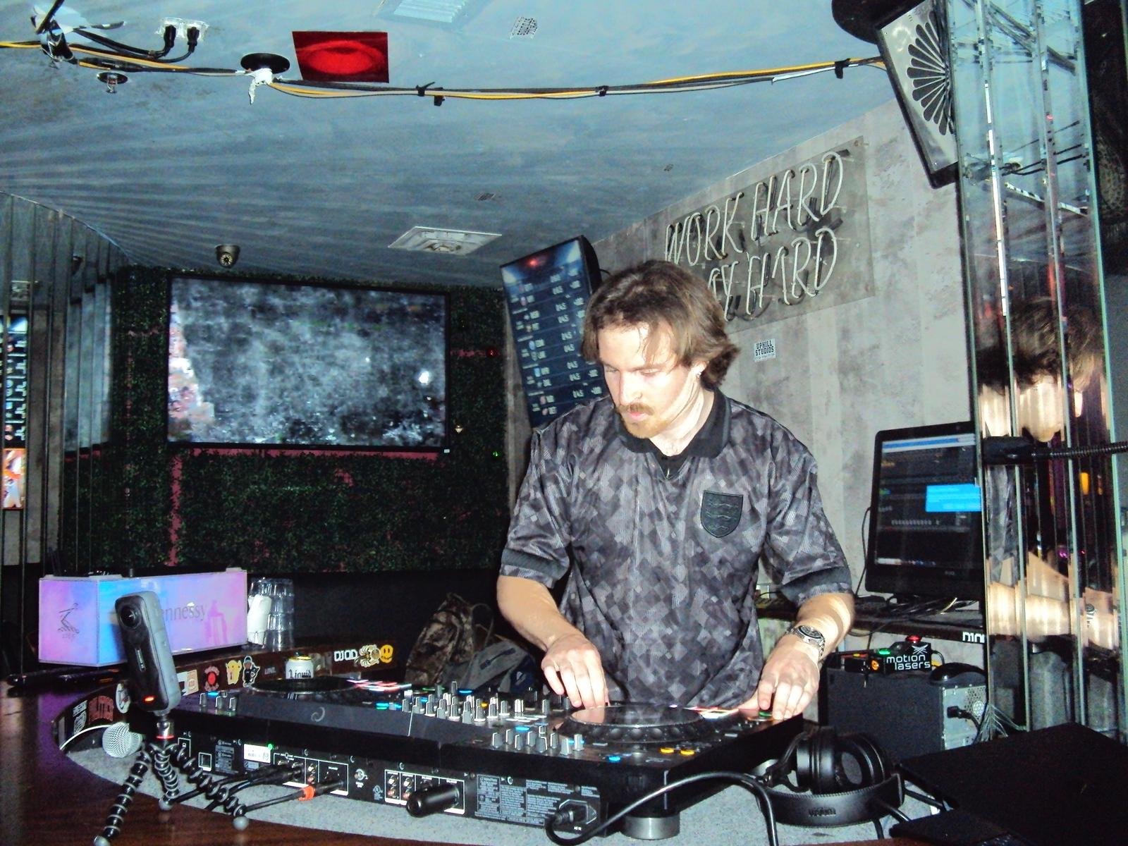DJ Sona performing at a nightclub with a large TV screen displaying visuals behind him. The wall has a sign with the phrase 'Work Hard Play Hard'.