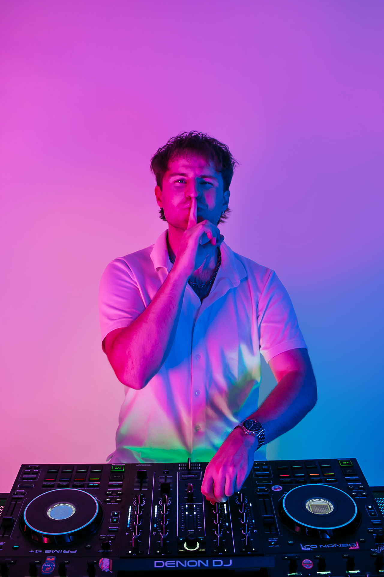 DJ Sona wearing a white shirt in a colorful, neon-lit room, operating a DJ controller and gesturing "shhh!".