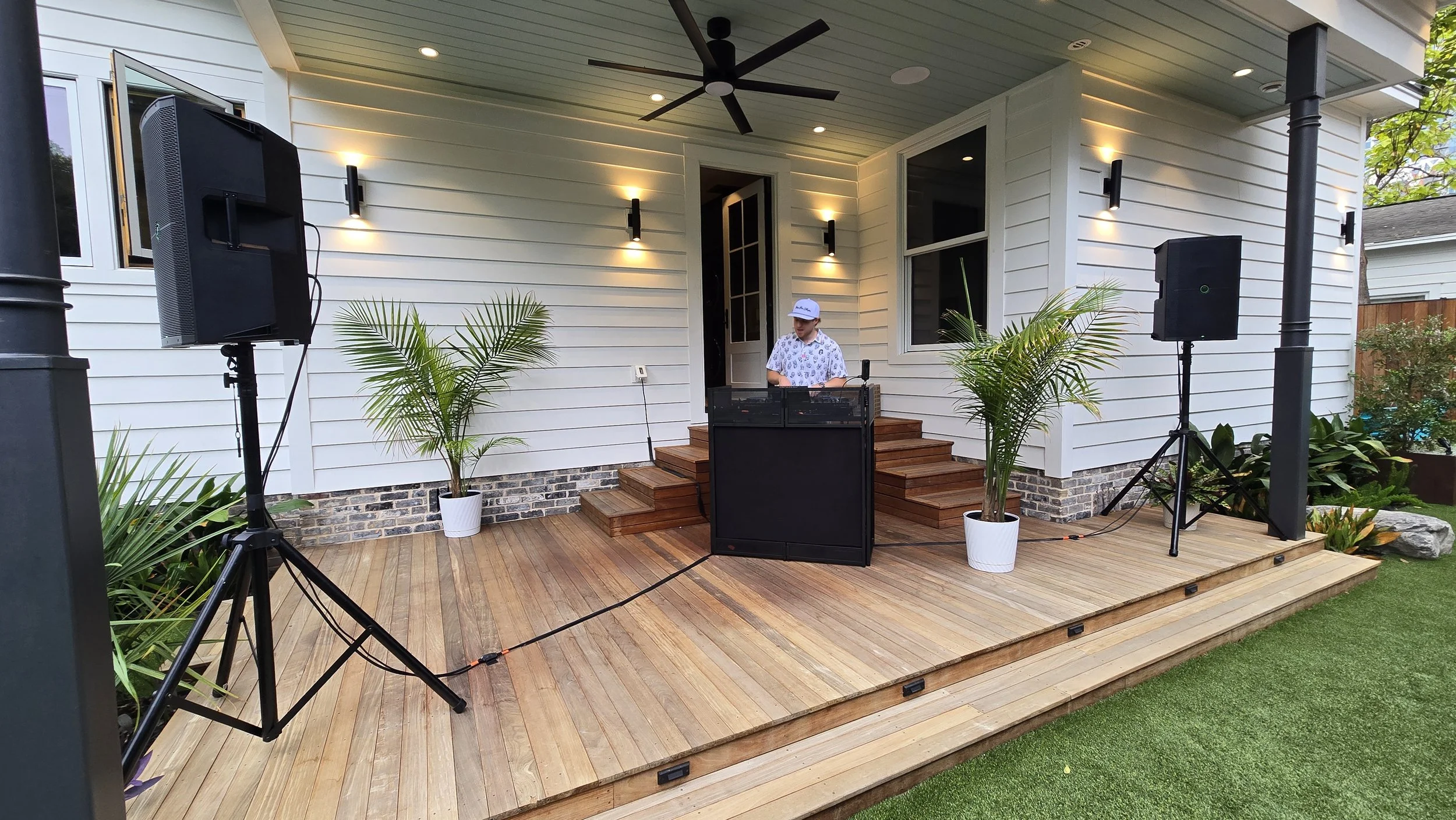 An outdoor backyard with a wooden deck. DJ Sona wearing a white hat and patterned shirt behind the DJ booth. There are two large speakers on stands, tropical plants, and modern wall-mounted lights on the house siding.