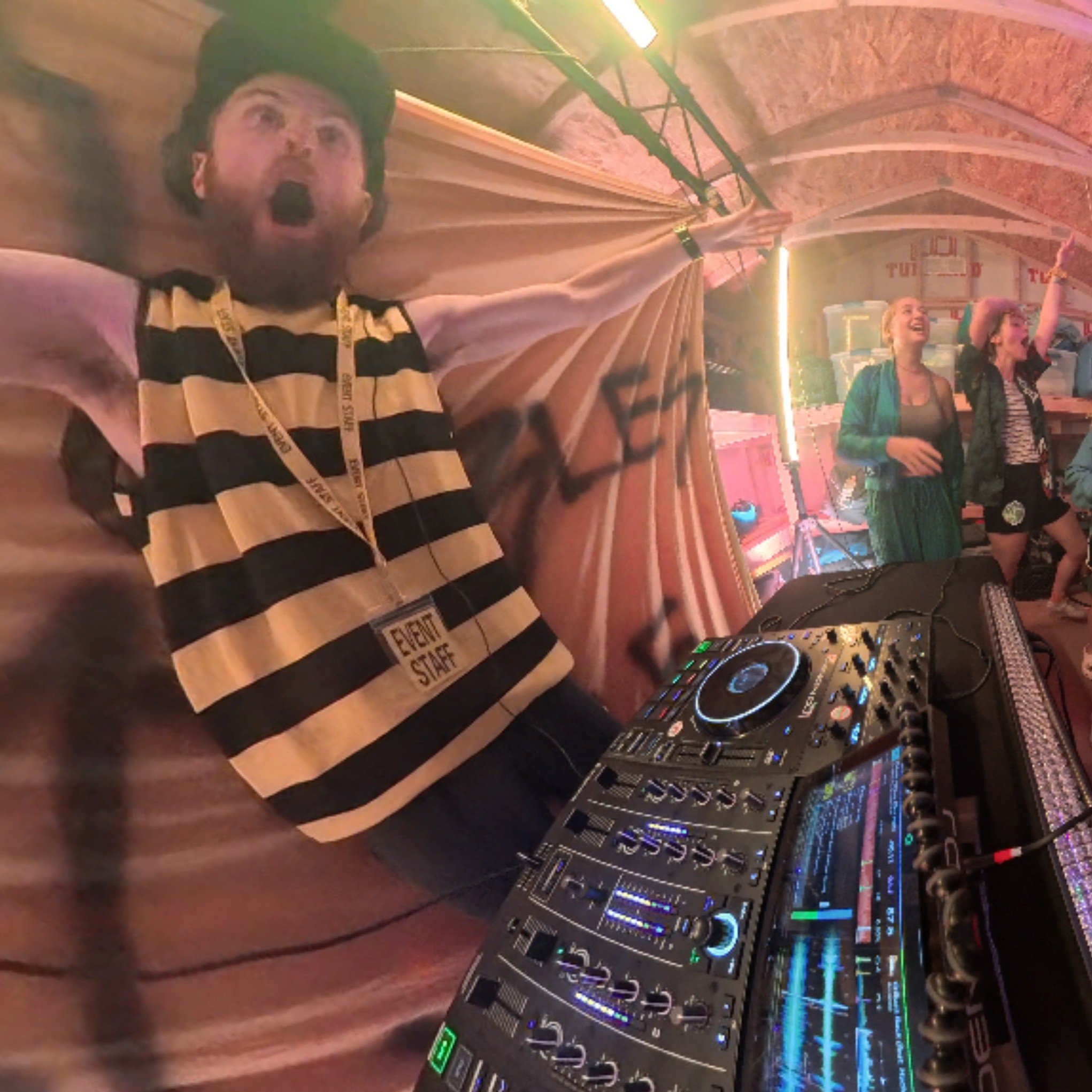 DJ Sona, dressed as a bumble bee, standing in front of DJ equipment with colorful lights. In the background, two women are smiling and raising their hands, inside a venue with wood-paneled arched ceilings and bright lights.