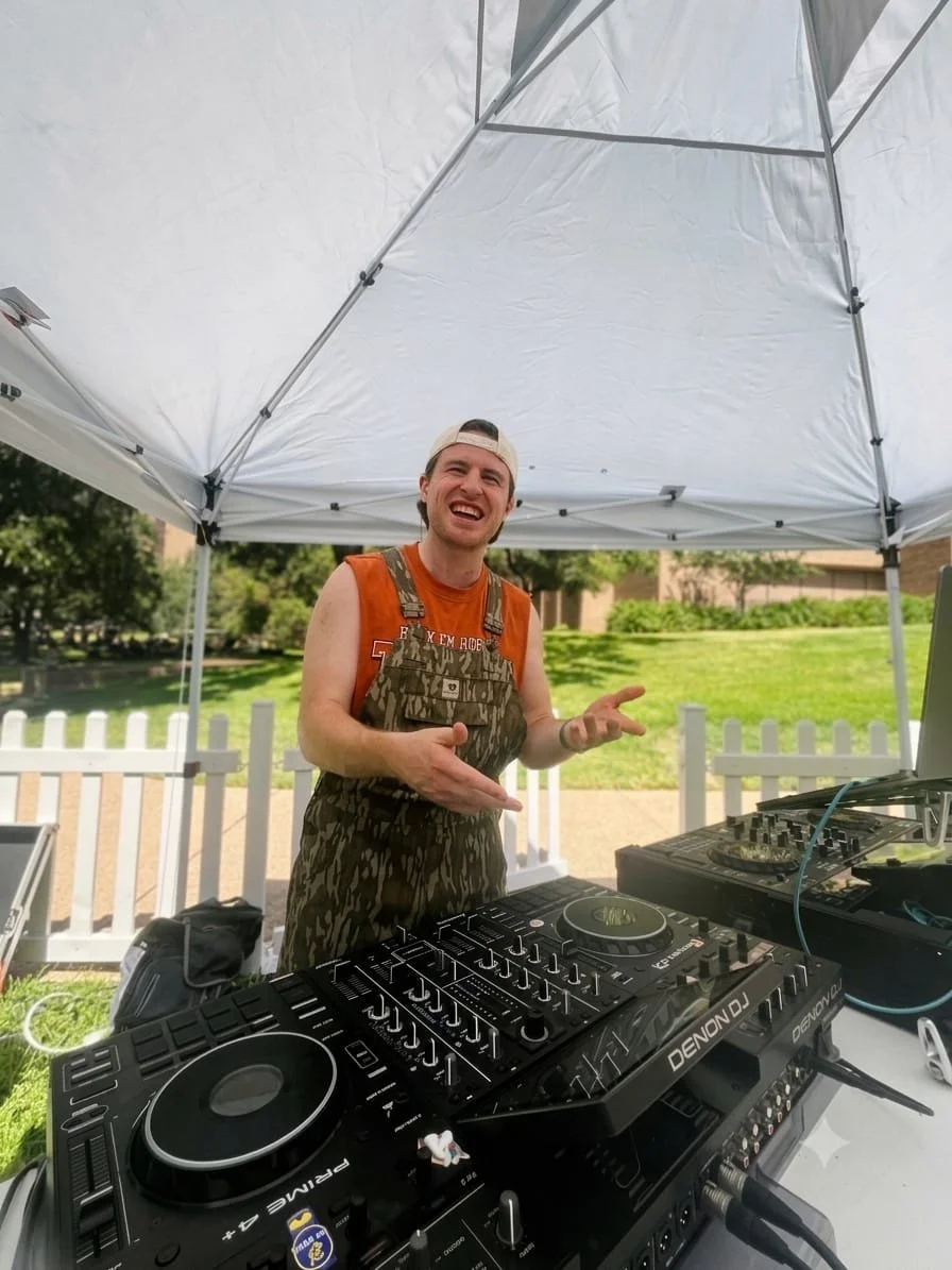 DJ Sona in camouflage overalls smiling under a white canopy outdoors with green grass and trees in the background at the UT Tailgate.