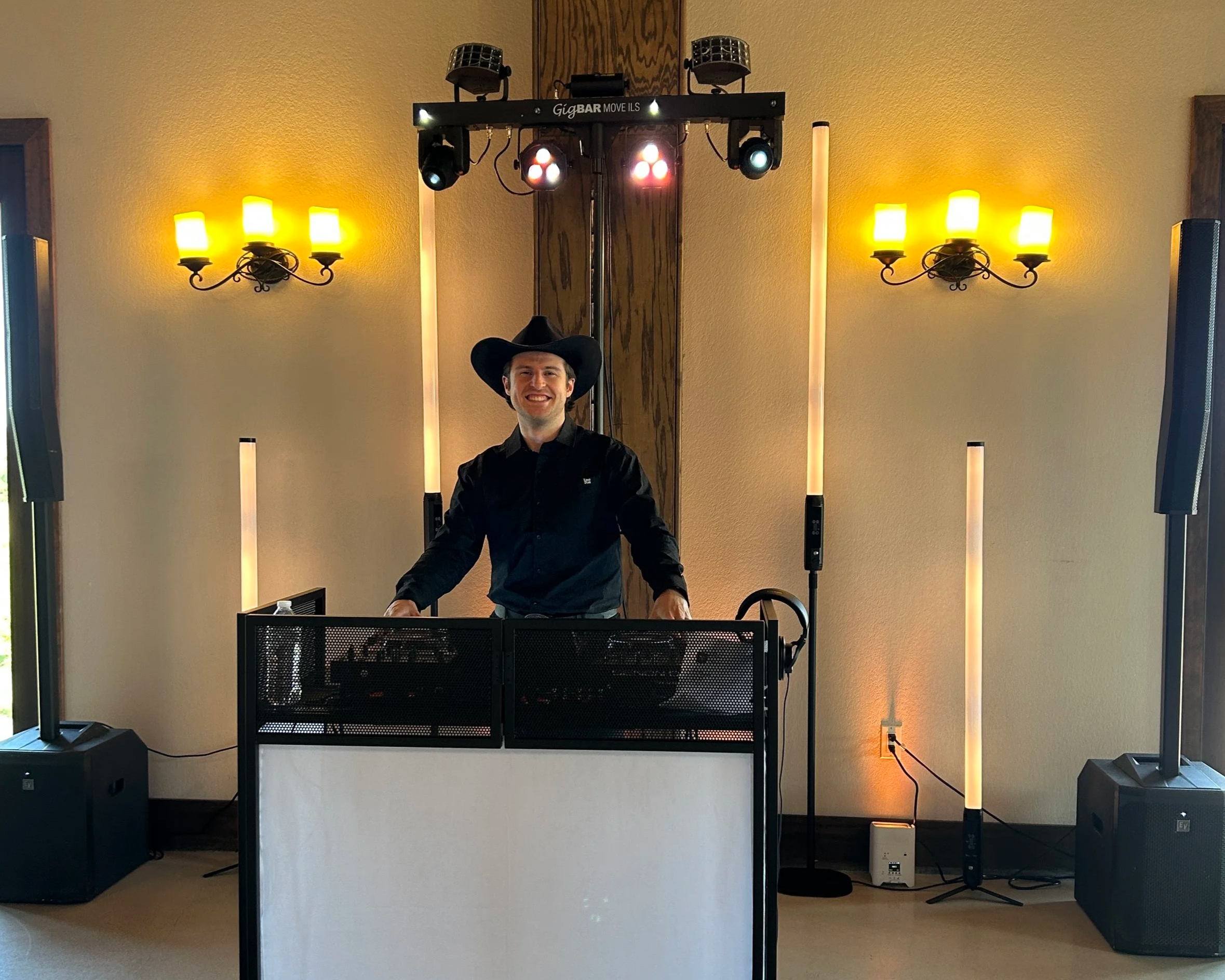 DJ Sona wearing a black cowboy hat and black shirt smiling while standing behind DJ equipment at an indoor wedding with yellow wall sconces and a DJ lighting rig overhead.