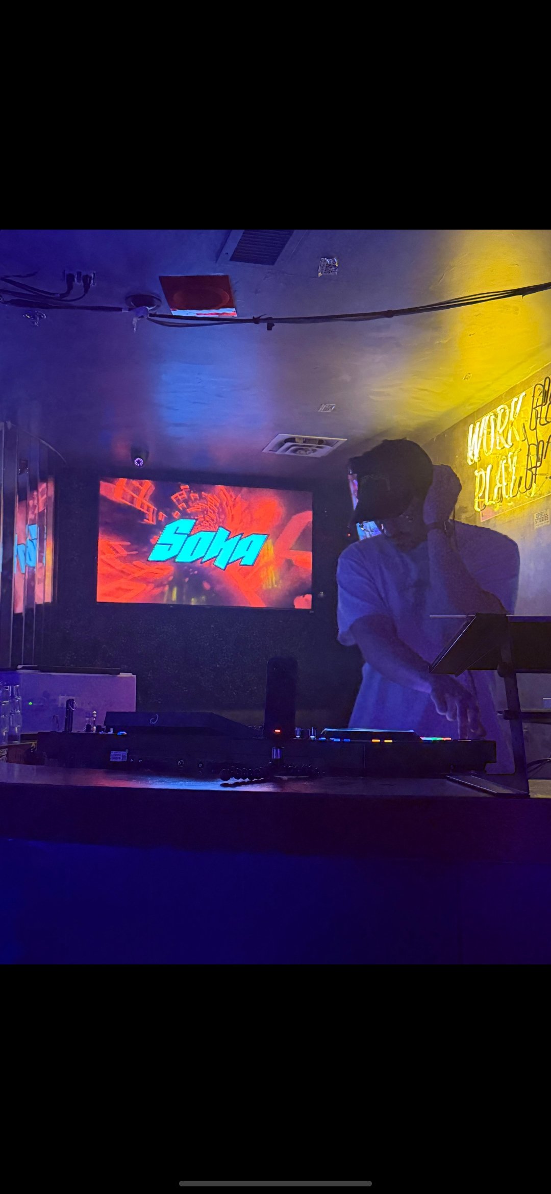 DJ Sona playing music at a club with a colorful screen displaying his name 'Sona' in the background. Neon sign that reads 'Work & Play' on the wall.