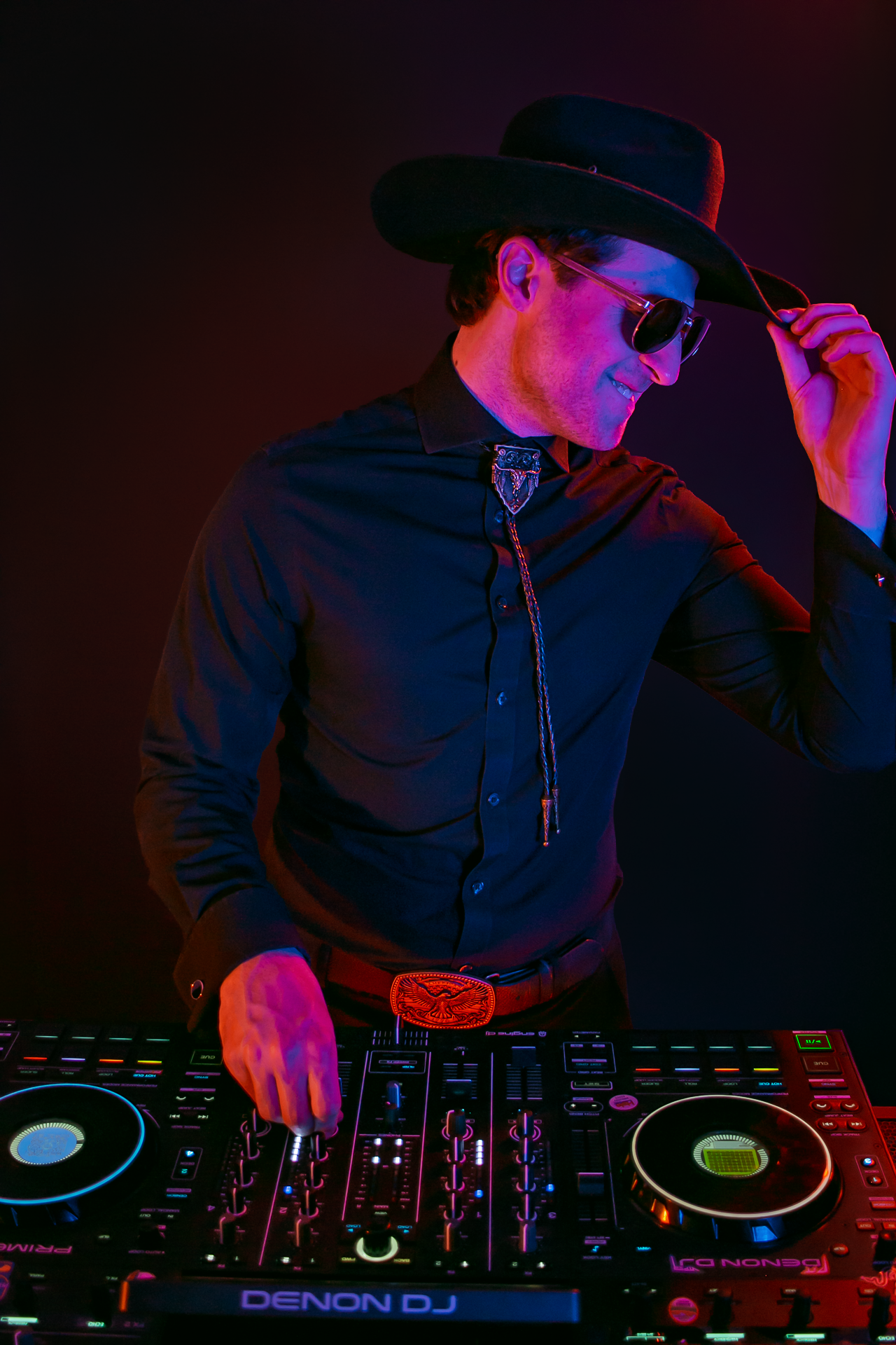 DJ Sona wearing a black cowboy hat, sunglasses, and a black shirt with an ornate bolo tie, standing behind a professional DJ controller with colorful lighting effects, set against a dark background.