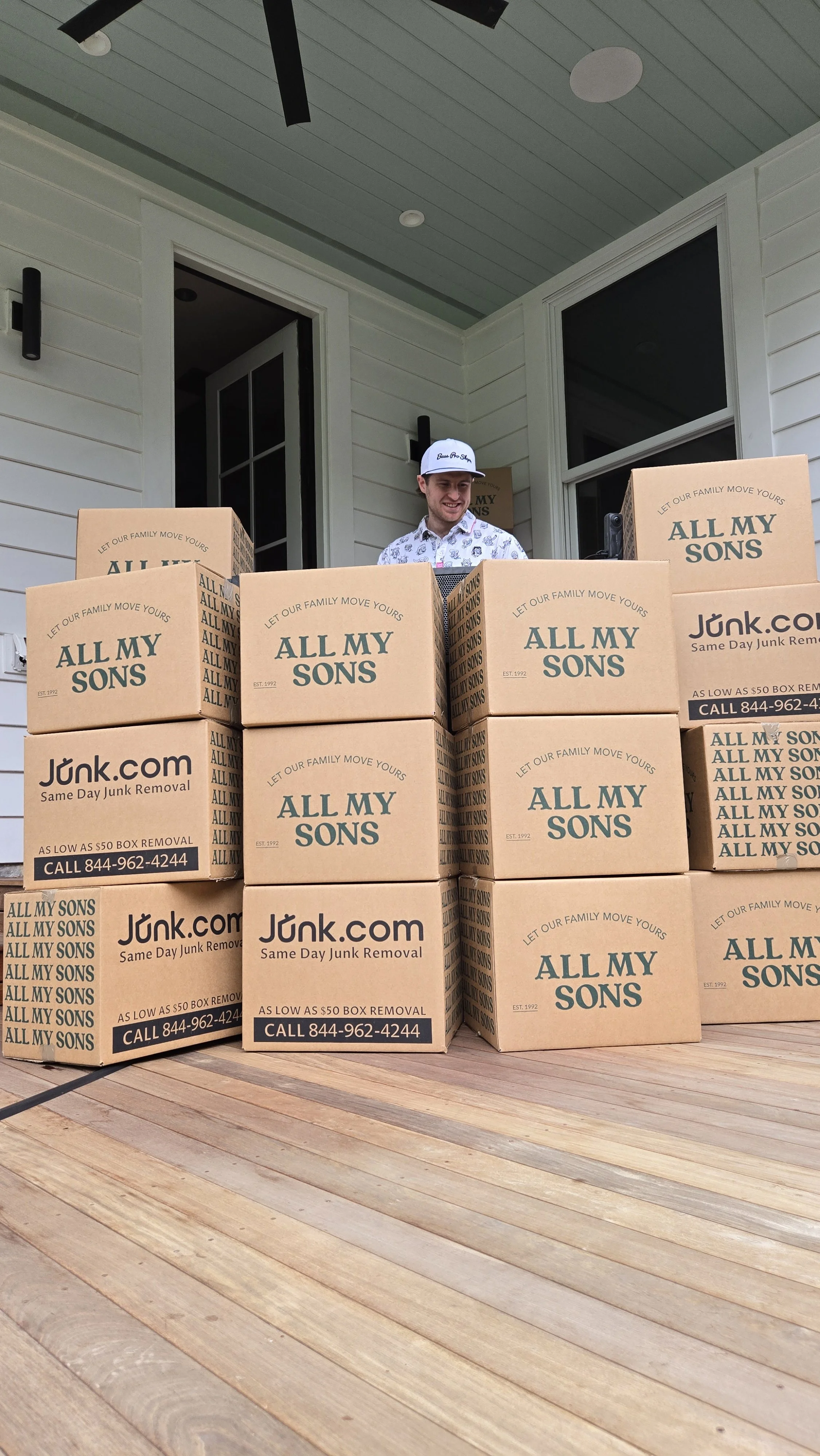 DJ Sona behind stacked cardboard boxes for the 'All My Sons' creator event on a wooden porch, with a house in the background.