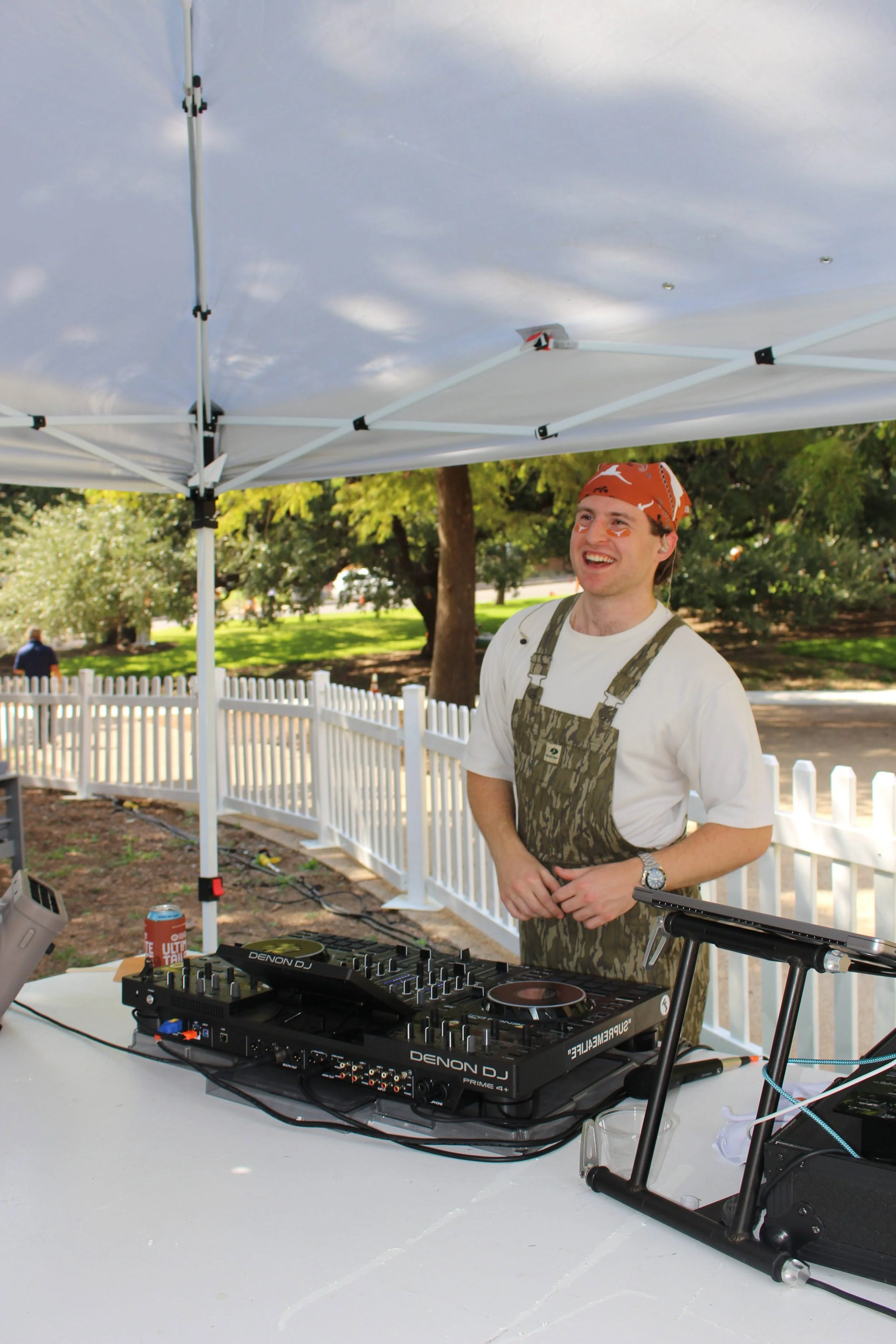 DJ Sona in a bandana, smiling, and dressed in a white shirt and camo apron, stands at a DJ setup outdoors under a white canopy. There is a white picket fence, trees, and a park-like setting in the background.