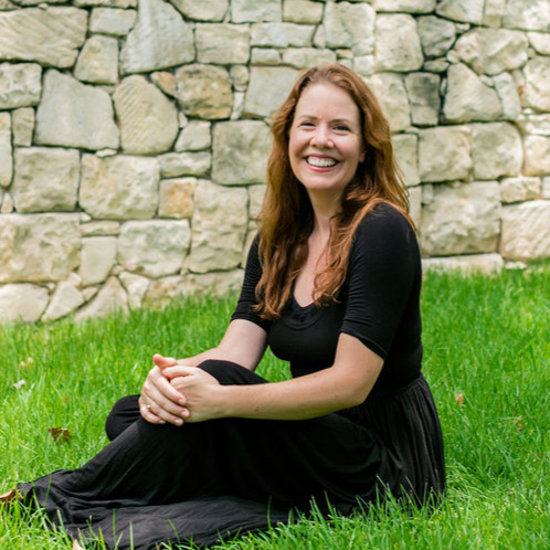 Laurel Crawford smiling outdoors, seated on grass in front of a stone wall.