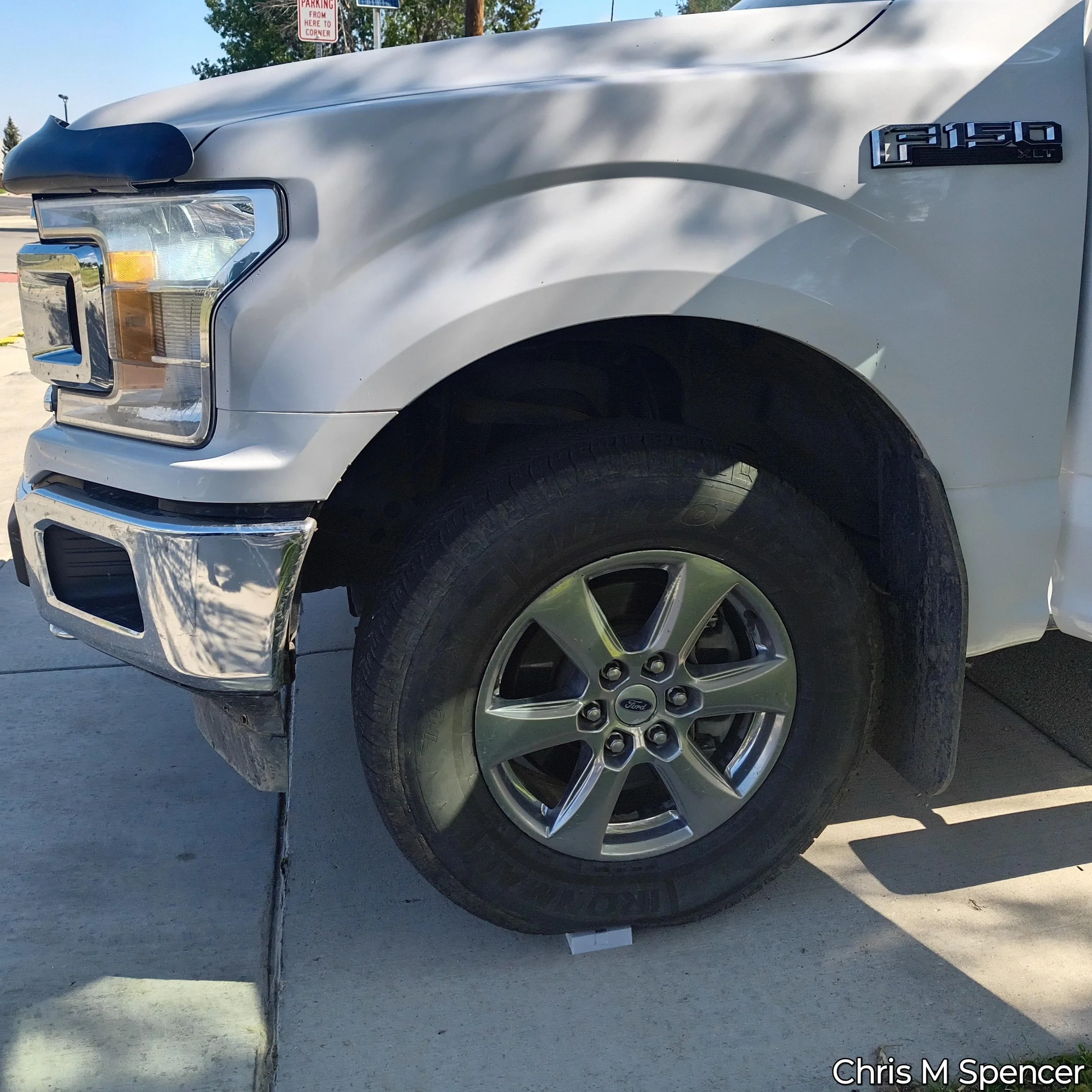 Close-up of a white Ford F-150 XLT pickup truck, showing the front right section including the headlight, grille, and wheel. The truck is parked outdoors, with parking signs and trees visible in the background. It is parked on top of one of razorcase
