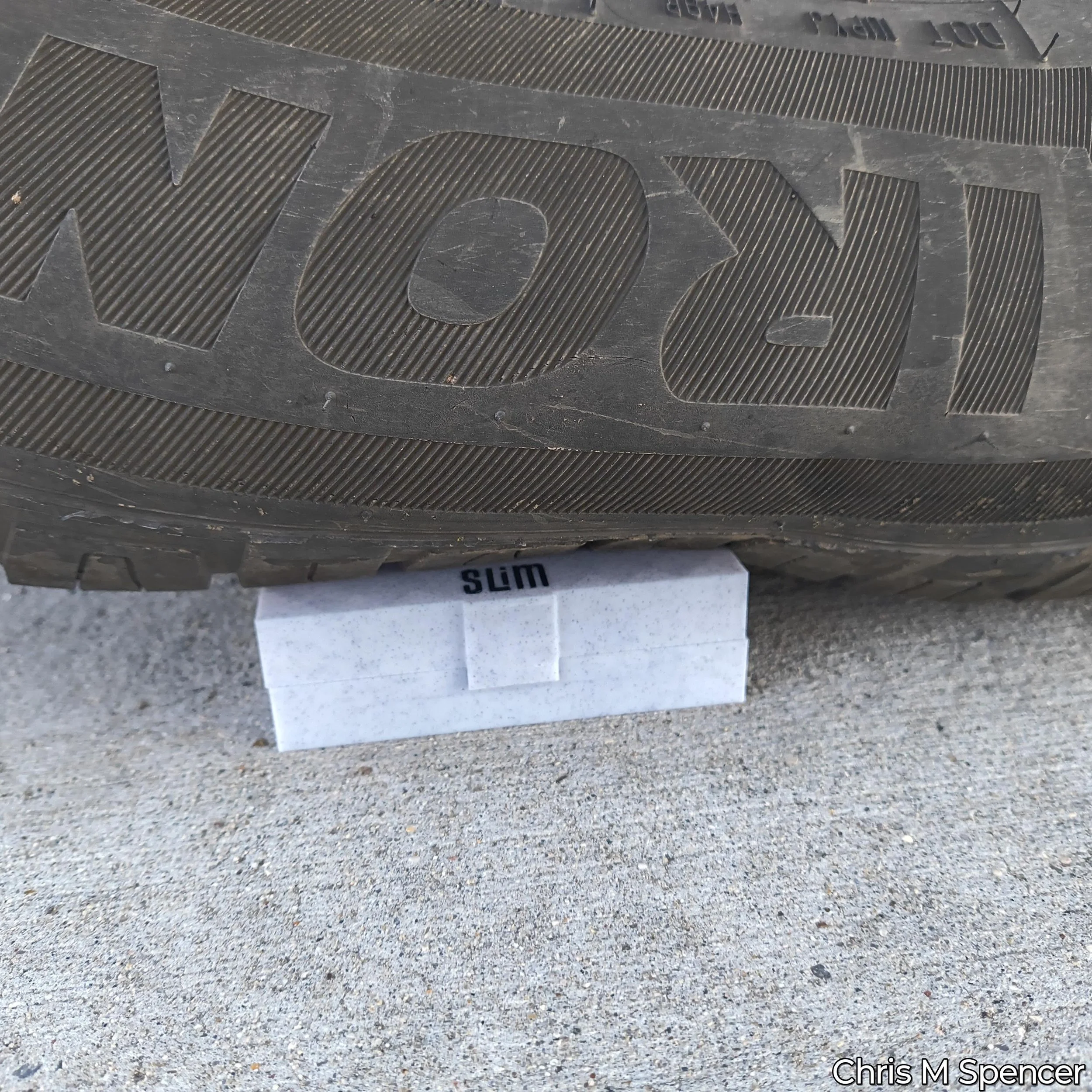 Close-up of a car's rubber tire with the word 'dyno' embossed in the tread pattern and a small section of the word 'slim' visible on a white surface beneath the tire, sitting on top of one of Chris M Spencer;s safety razor cases to show the durabilit