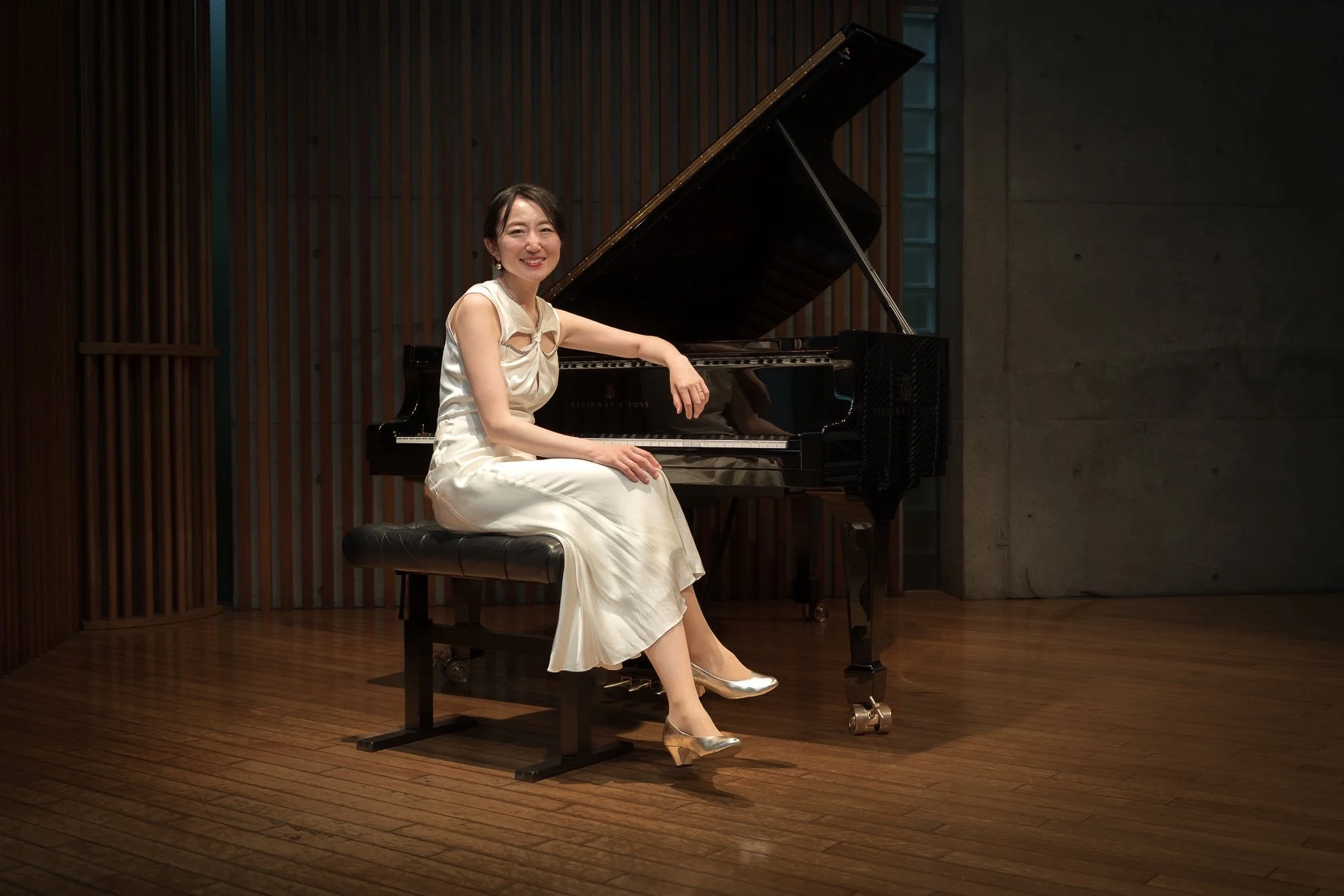 A woman dressed in a white gown sitting beside a grand piano on a wooden stage, smiling at the camera.
