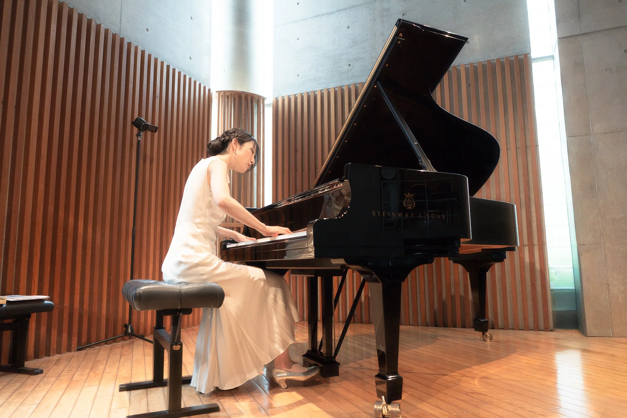 A woman in a formal white dress playing a black Steinway & Sons grand piano in a modern concert hall with wooden paneling and concrete walls.